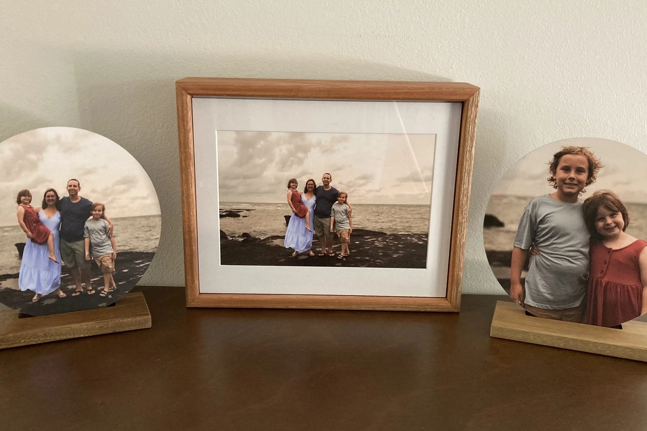 Family photo with four people, two adults and two children, on a beach, displayed in a wooden frame and two circular photo prints on wooden stands.