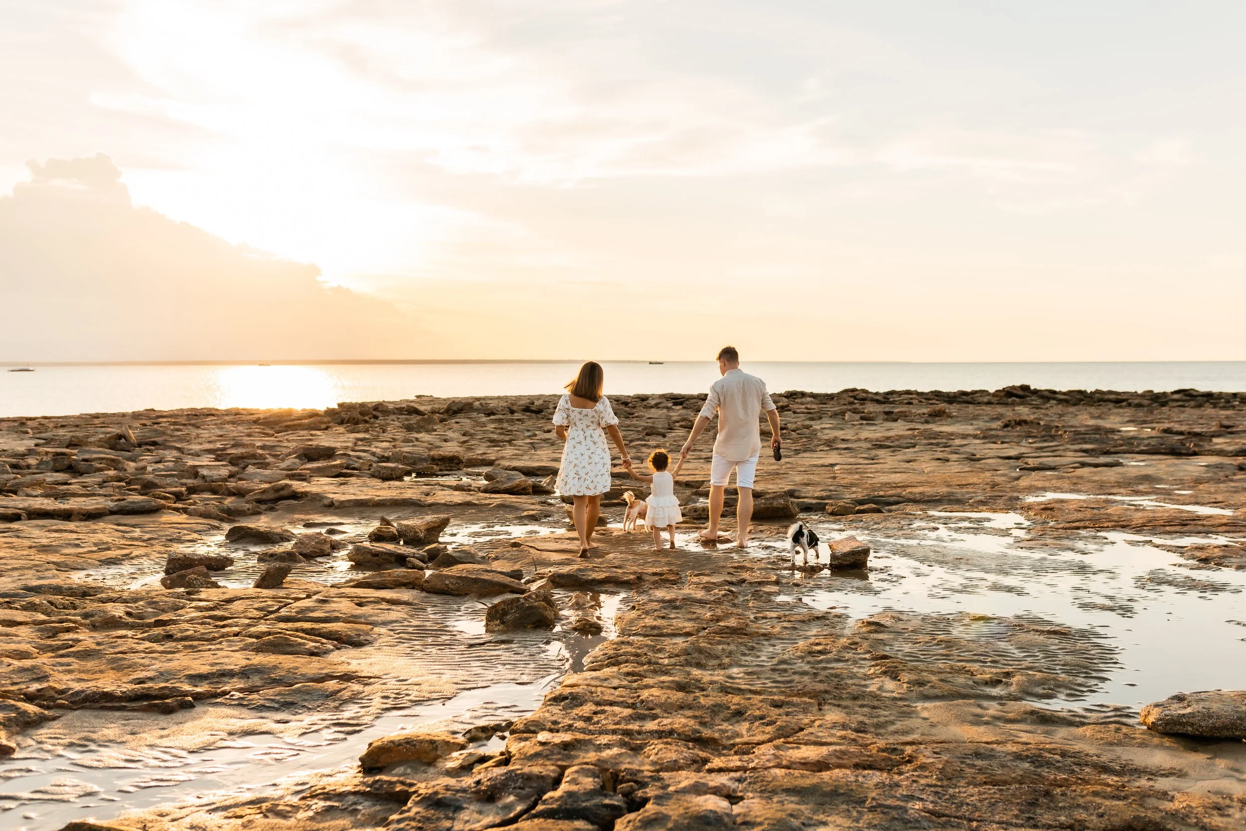A family of four, including two children, walks along a rocky shoreline at sunset, with two dogs. The sky is partly cloudy with the sun low on the horizon.