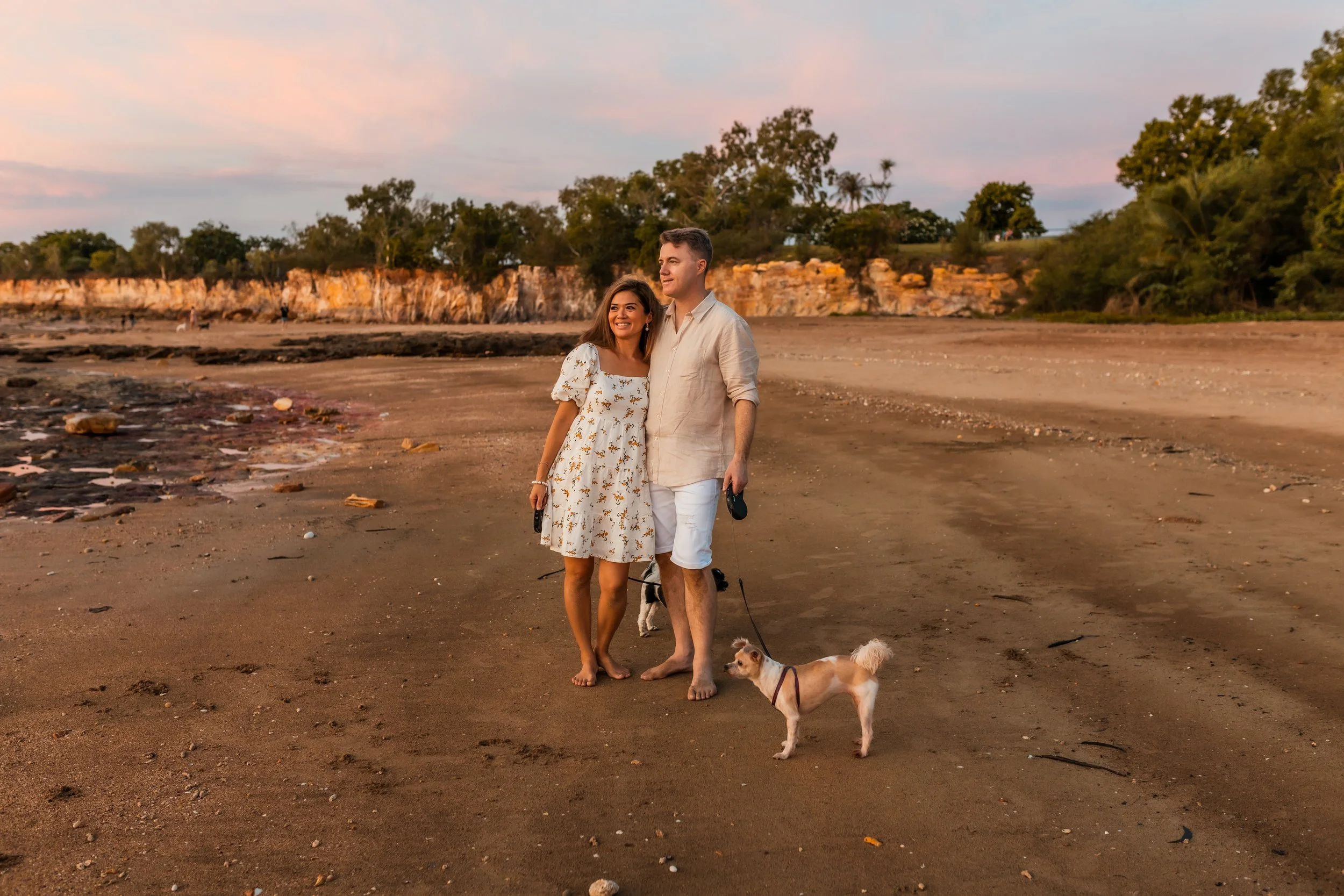 A happy couple walking on a sandy beach at sunset, with their dog. The woman is wearing a white floral dress, and the man is wearing a light-colored shirt and white shorts. There are trees and rocky cliffs in the background.