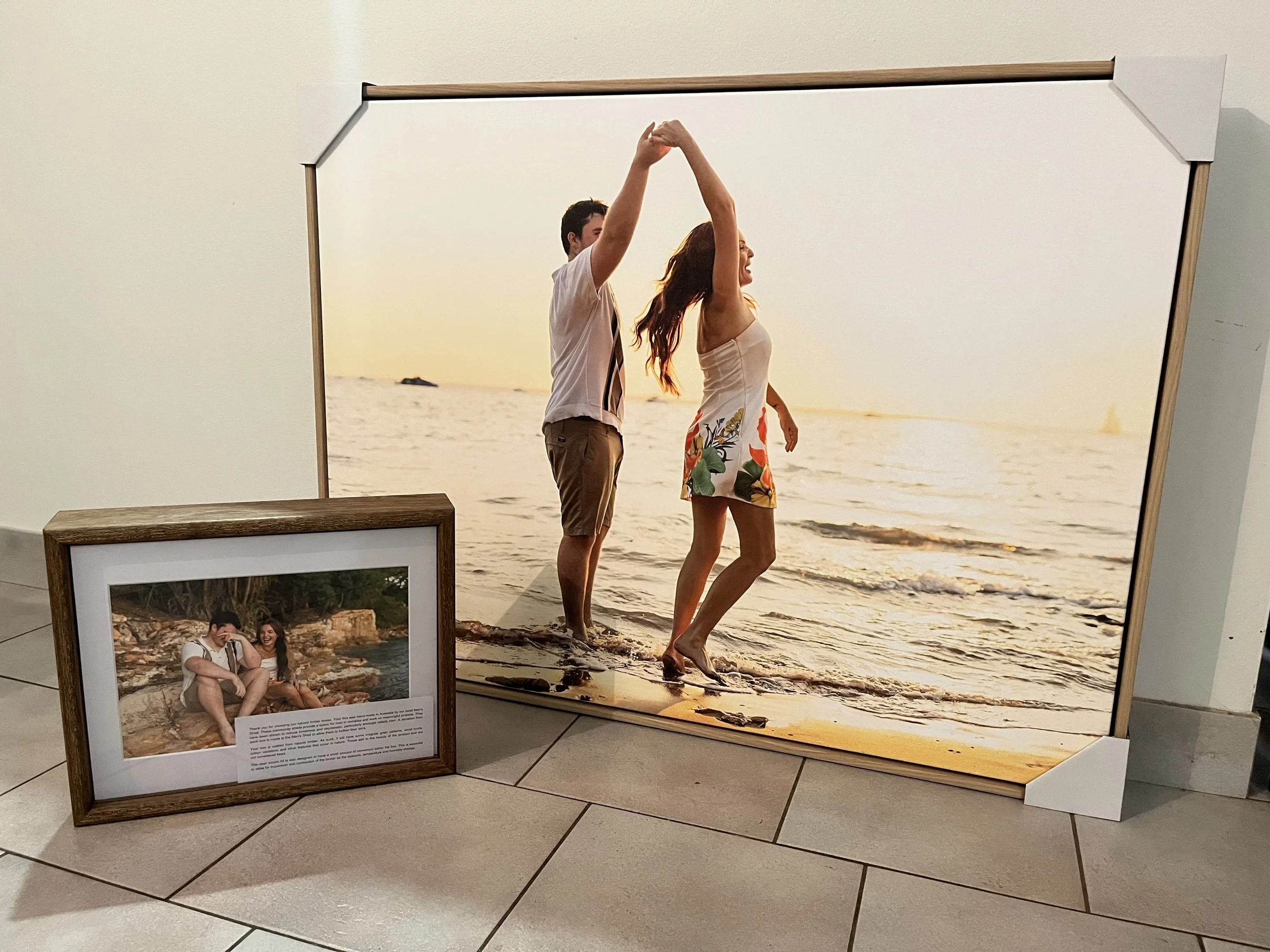 Two people dancing on the beach during sunset, with a smaller framed photo of a couple sitting by a river placed on the floor nearby.