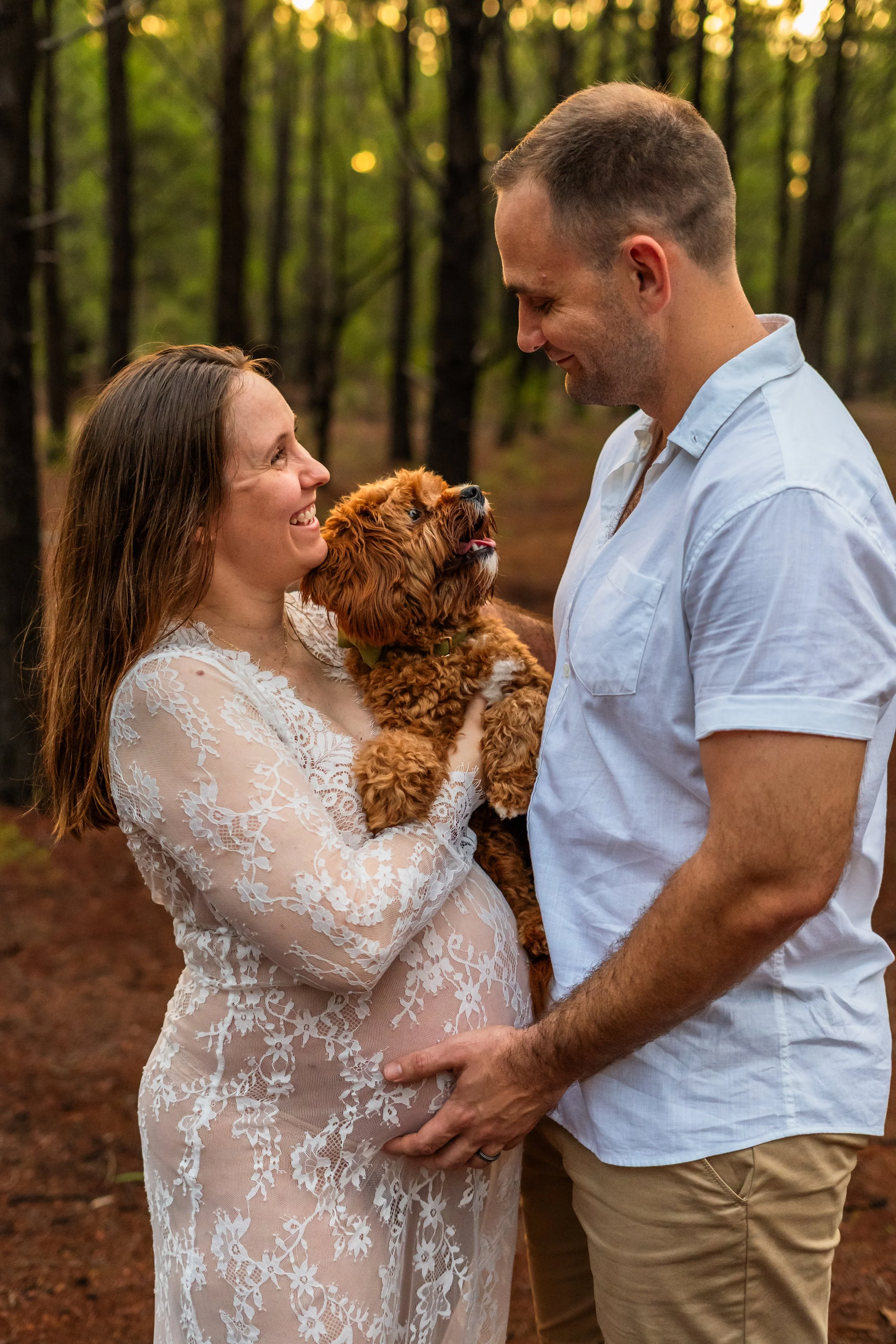 A pregnant woman and a man standing in a forest, smiling at each other, holding a small brown dog together.