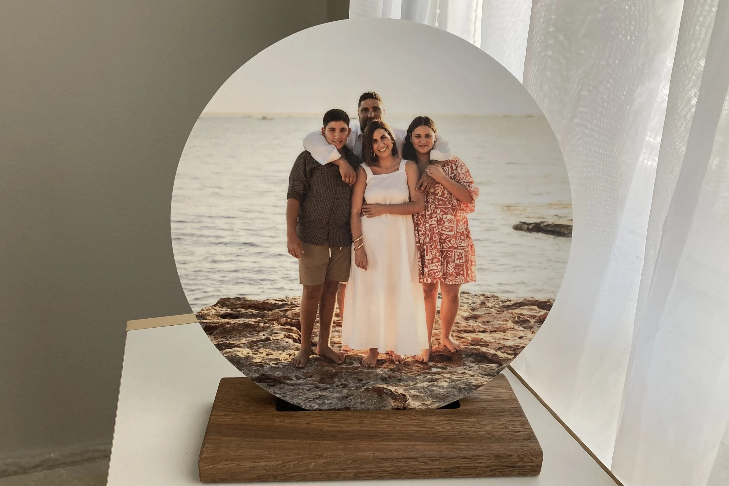 Photo on a round wooden stand of a family of five outdoors at the beach during sunset, standing on rocks near the water, smiling and posing for the camera.
