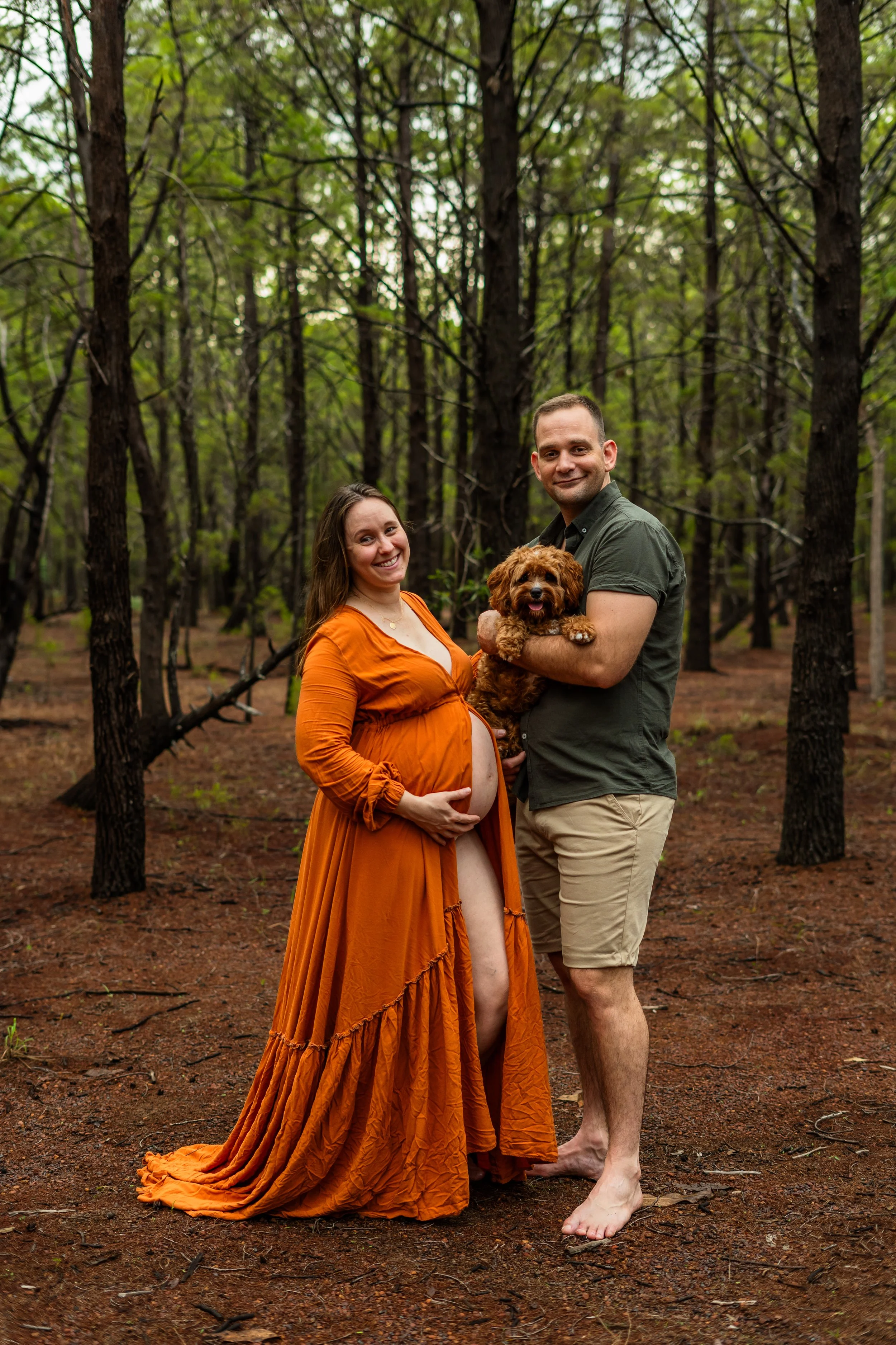 An expecting woman in an orange dress and a man holding a small brown dog, standing barefoot in a forested area with tall trees and a dirt ground.