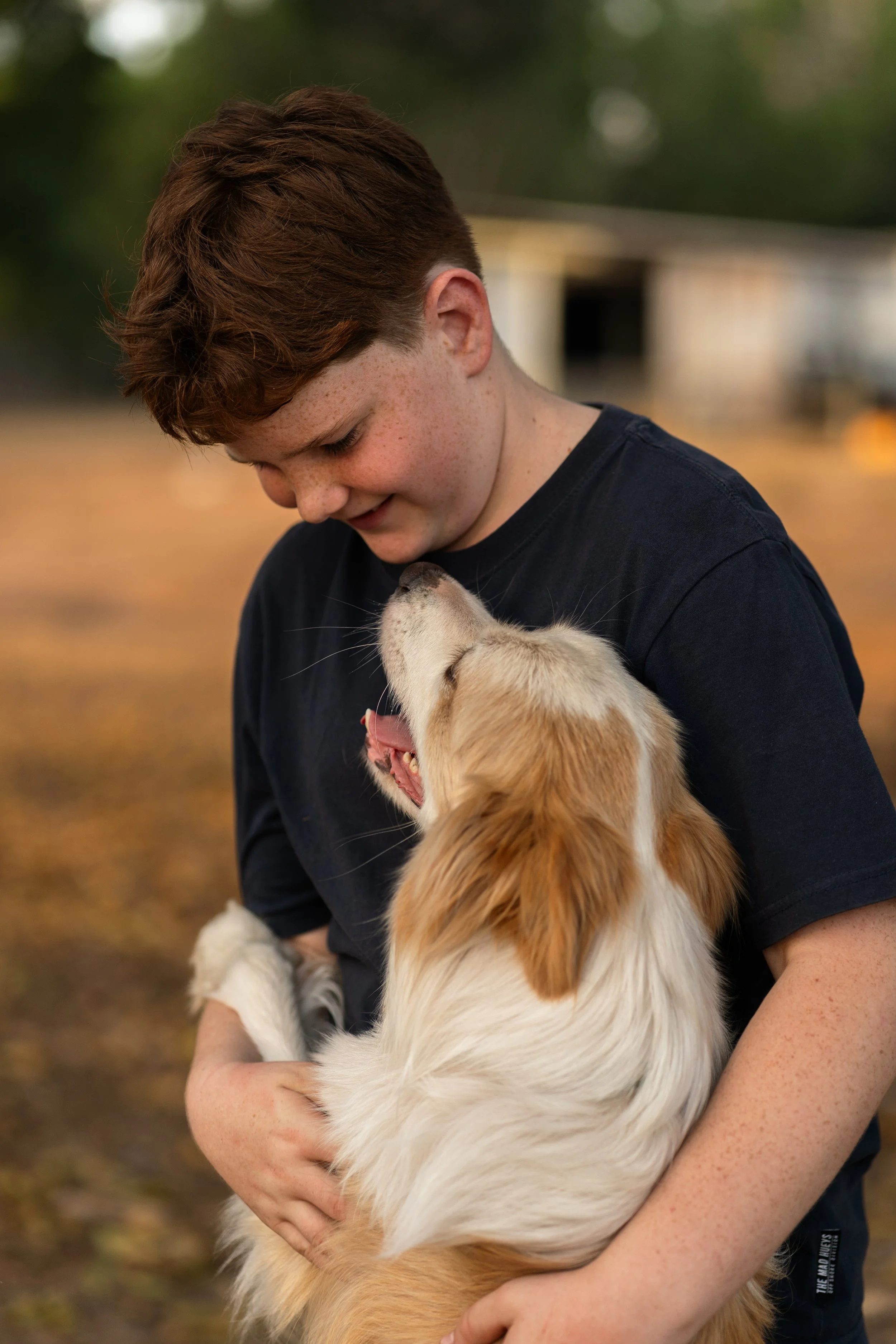A young boy with red hair and freckles holding and cuddling a golden retriever dog outside during the daytime.