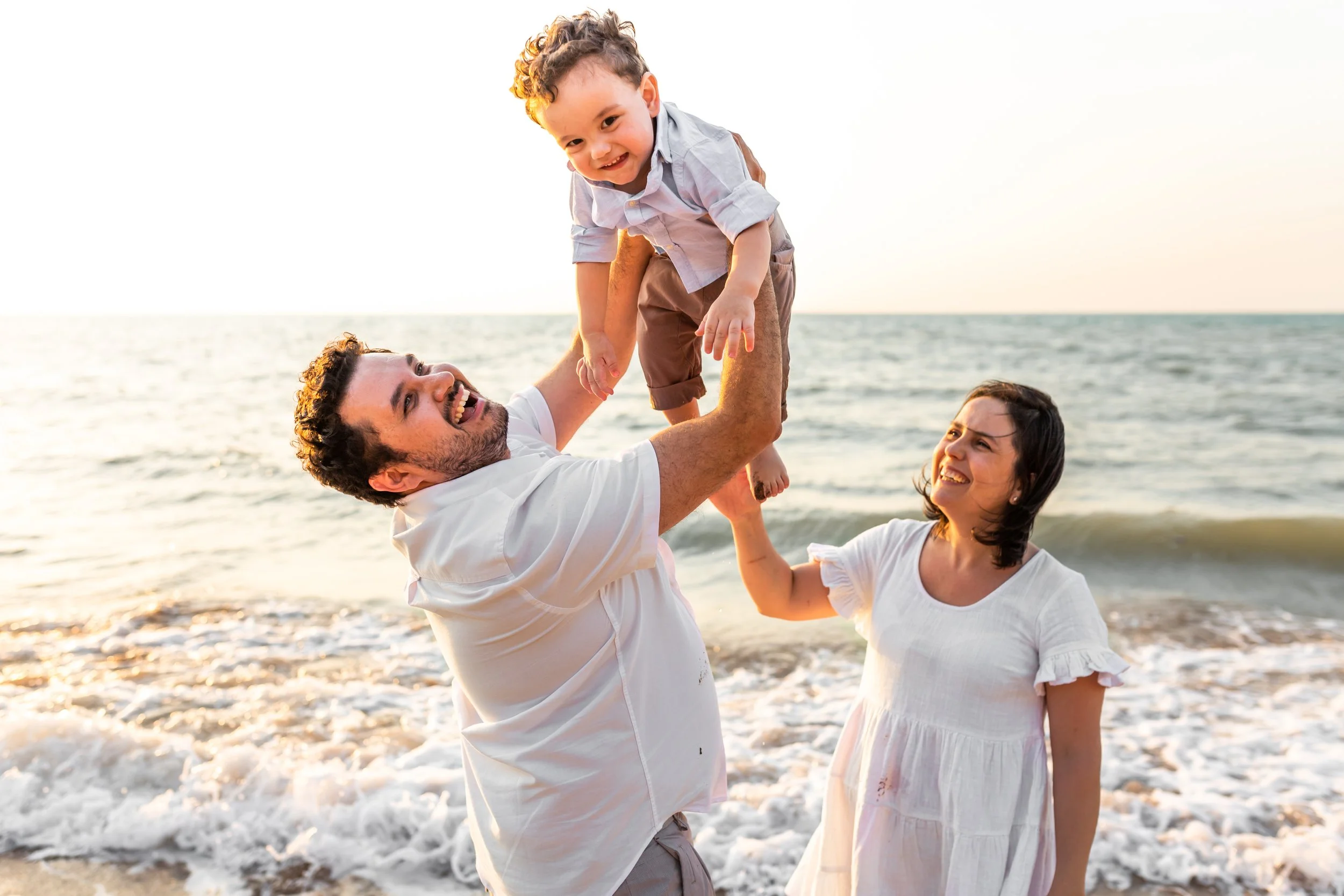 A family of three enjoying time at the beach during sunset. The father is lifting a young boy, and the mother is smiling at them.