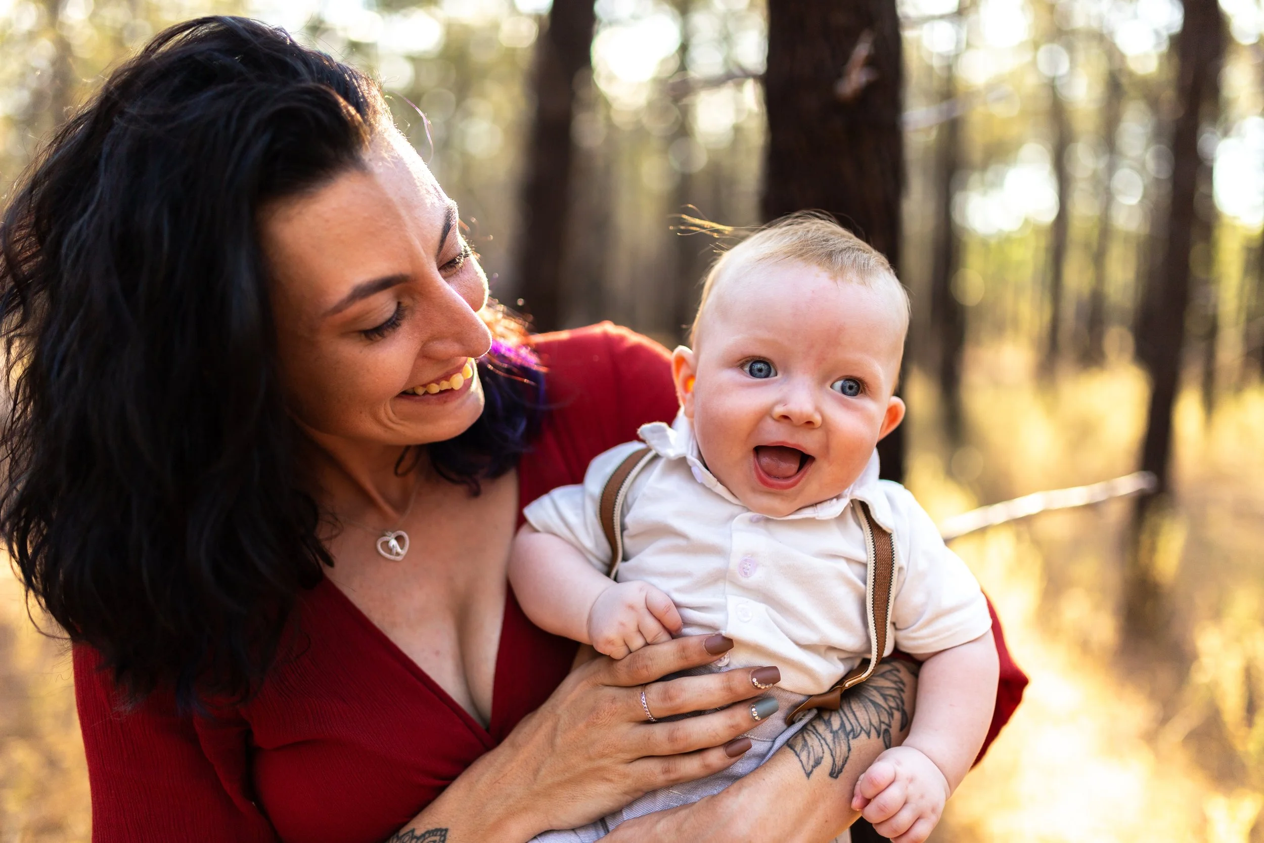 A woman with dark hair and a red top holding a smiling baby with blue eyes in a forest during daytime.