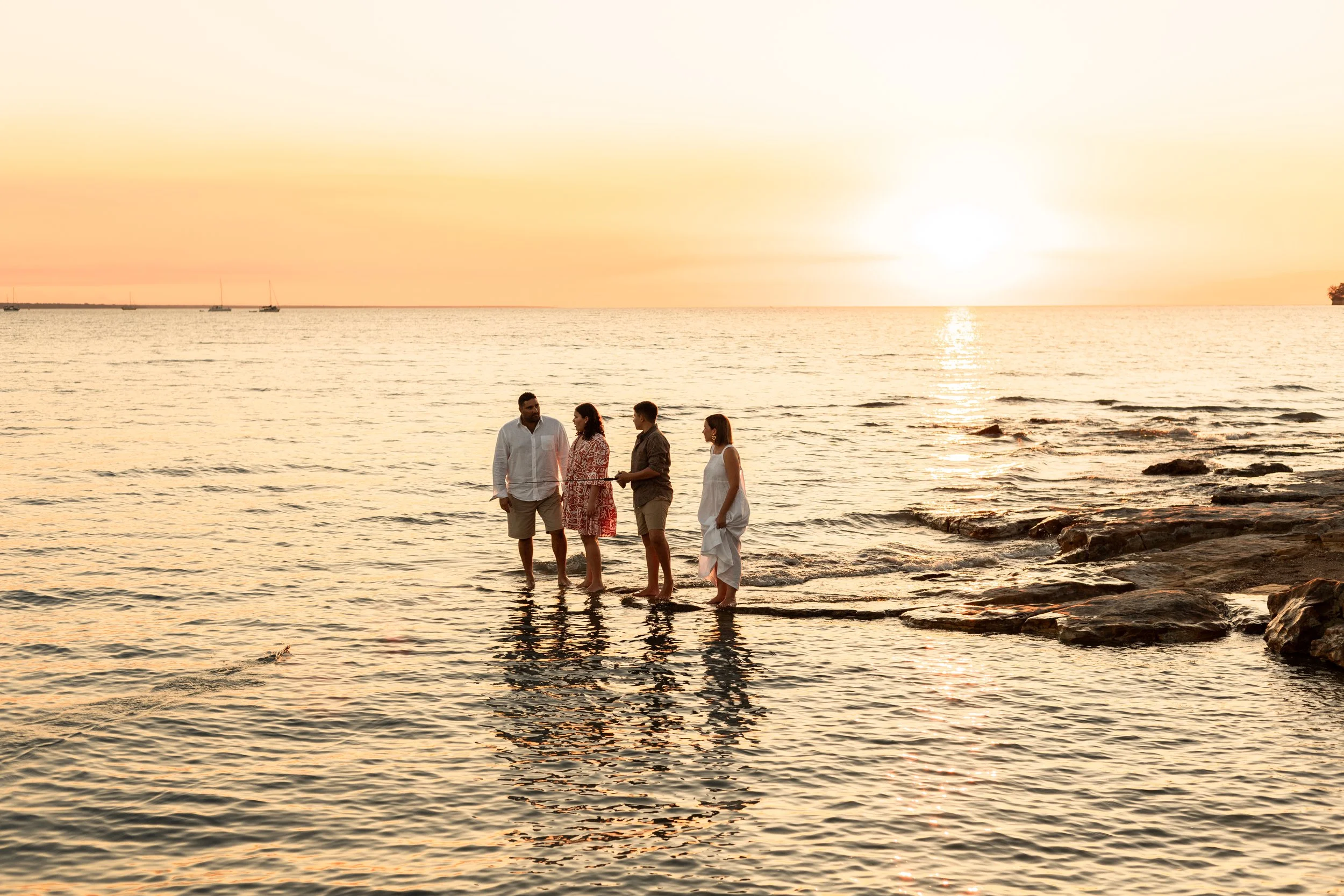 Four people standing in shallow ocean water at sunset, with sailboats and a distant shoreline in the background.