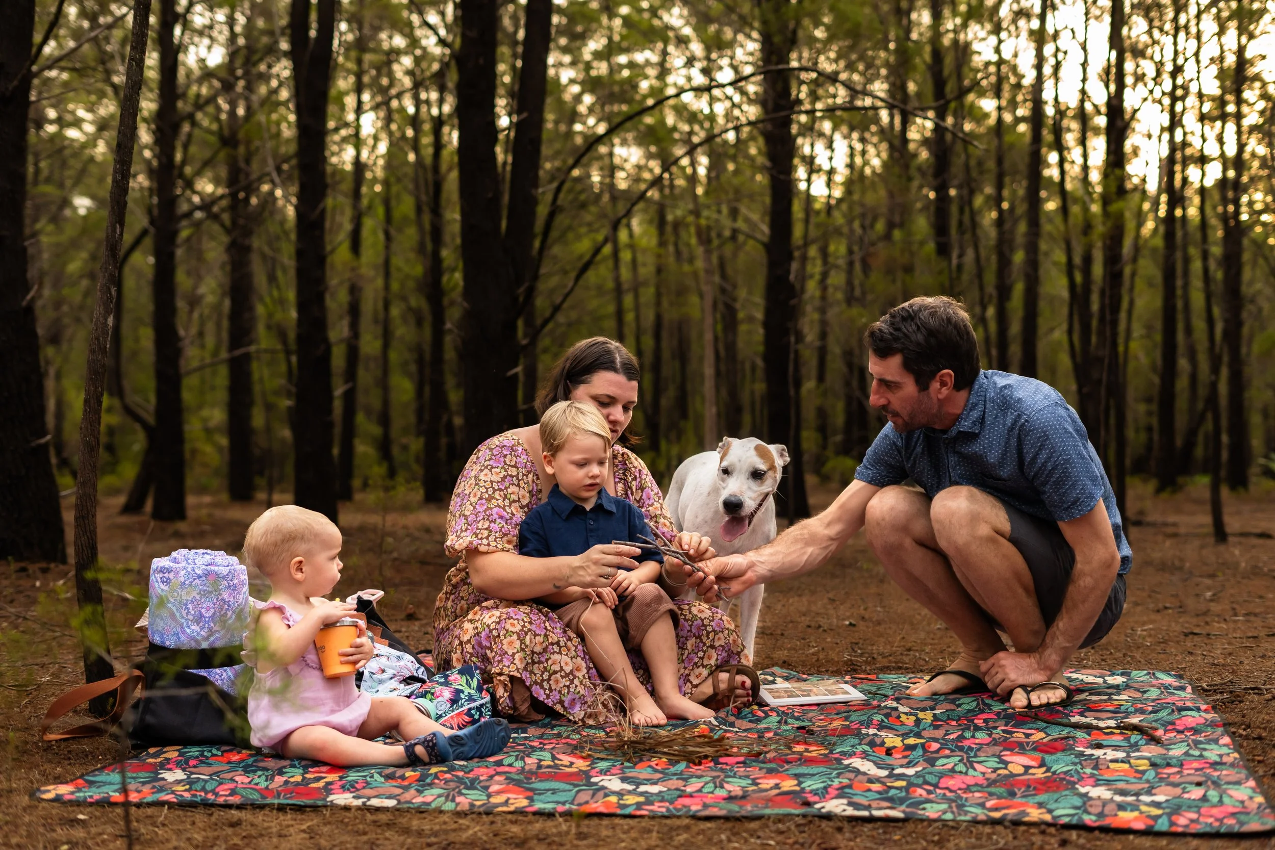 Family sitting on a colorful blanket in a forested area during sunset, with two adults, three children, and a dog.