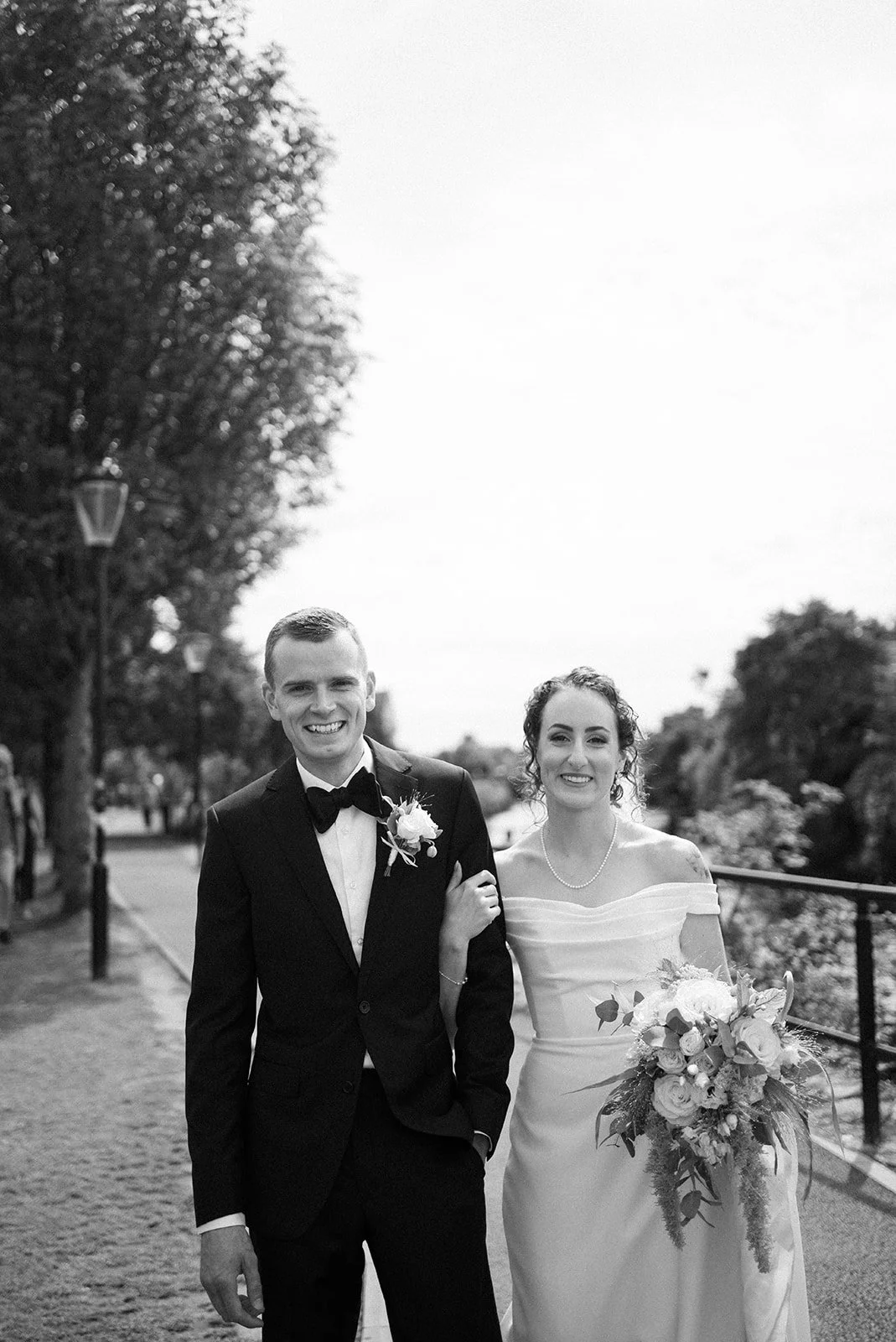 A smiling bride and groom in a black and white photograph, standing outdoors on a pathway lined with trees and lampposts. The bride is wearing a white wedding dress and holding a bouquet, while the groom is in a suit with a bow tie and boutonniere.