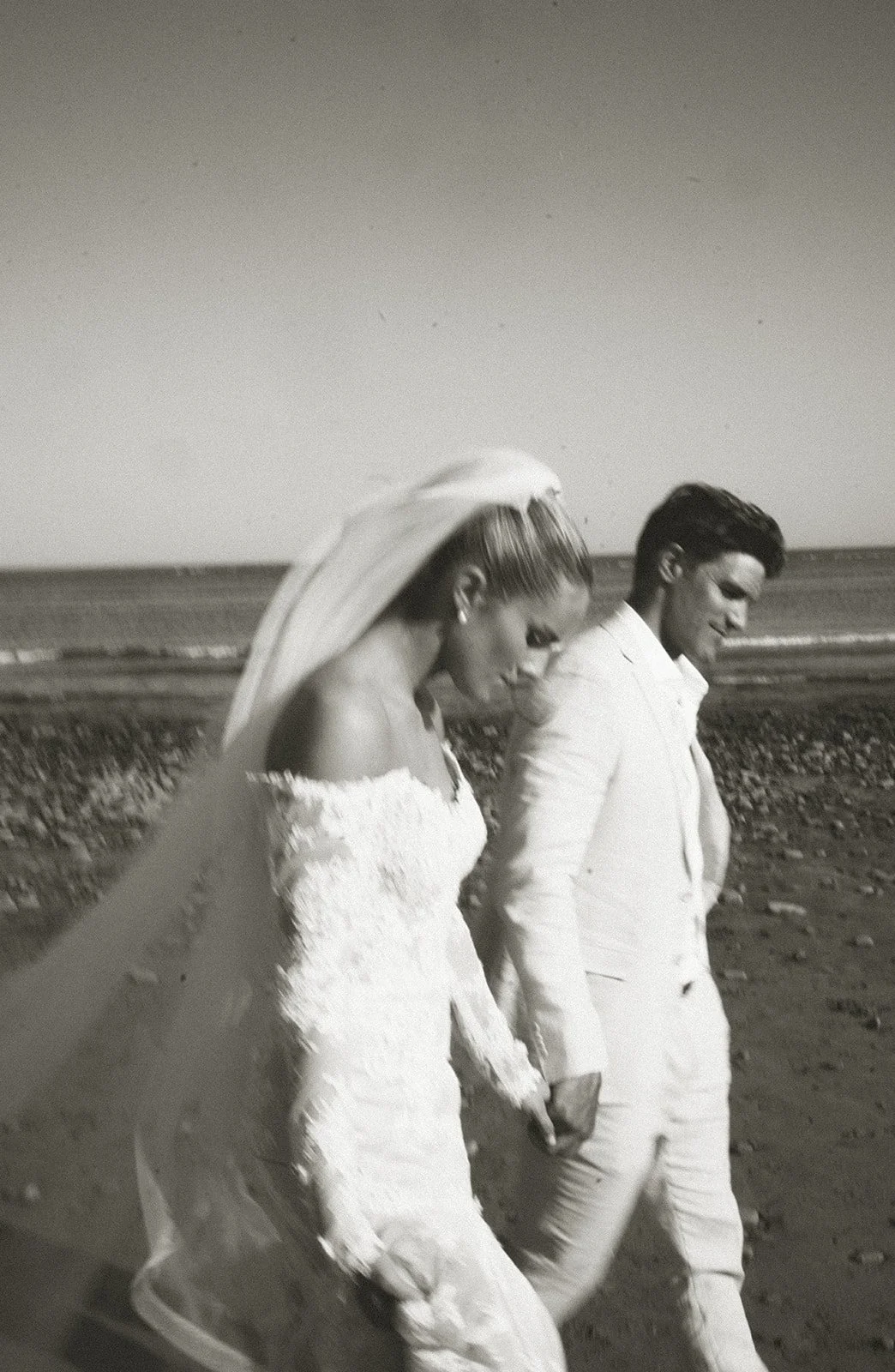 Bride and groom walking on a beach, wearing wedding attire, holding hands in a monochrome photograph.