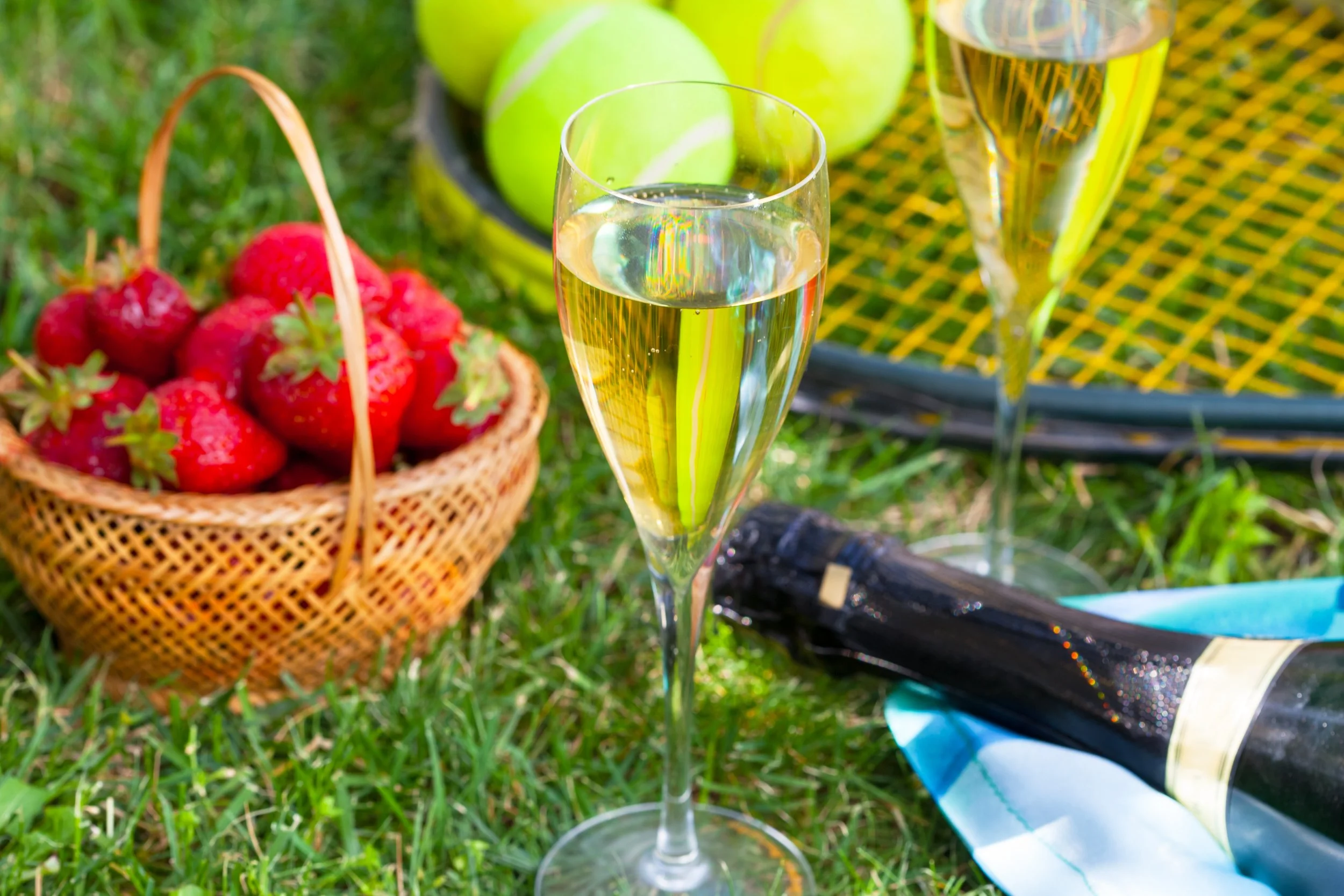 Champagne glasses, basket of strawberries, tennis racket, tennis balls on grass.