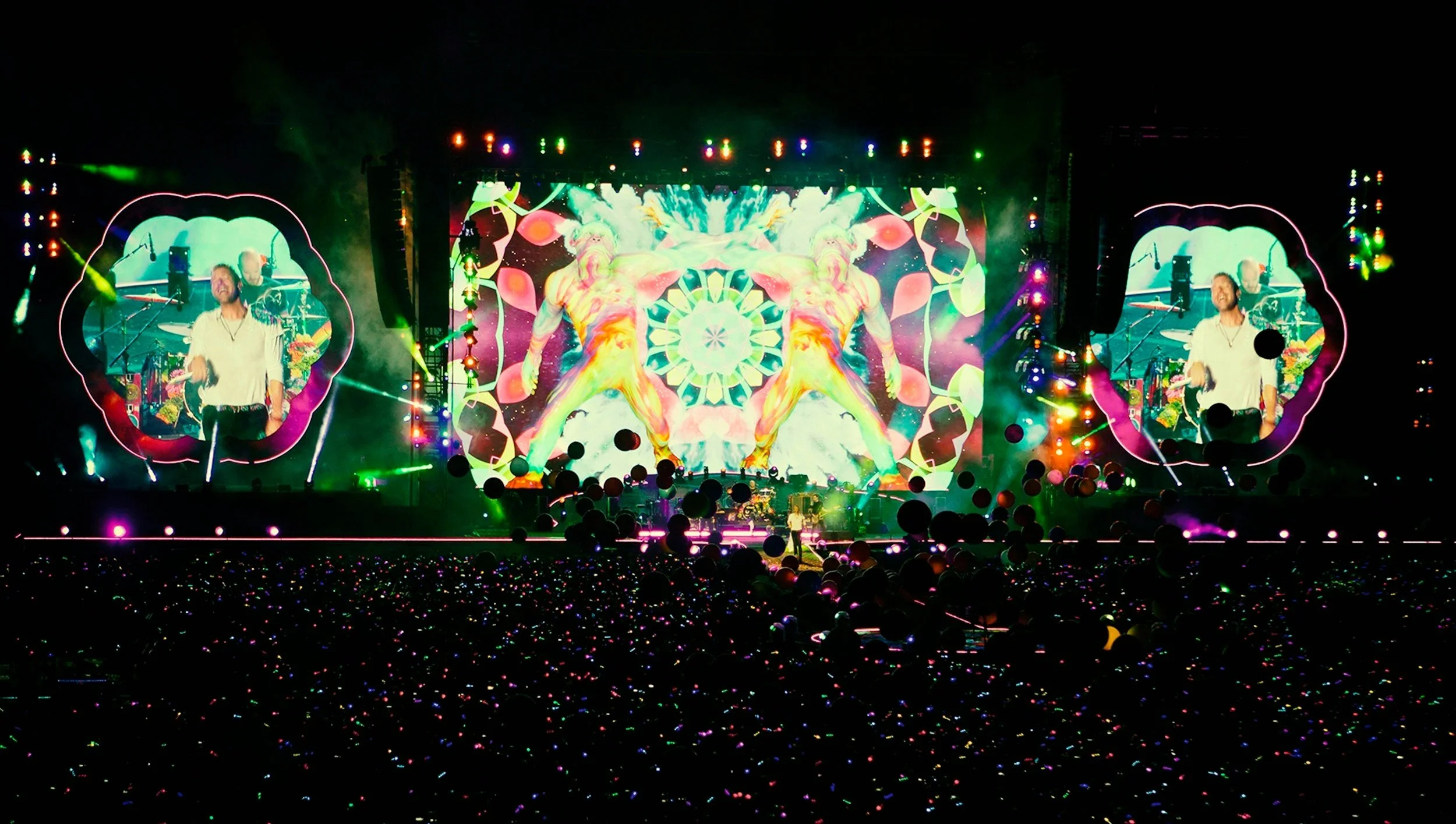 Coldplay performing on xoncert stage with colorful lighting and video screens showing abstract visuals. A large crowd is watching, with colorful lights visible among them.