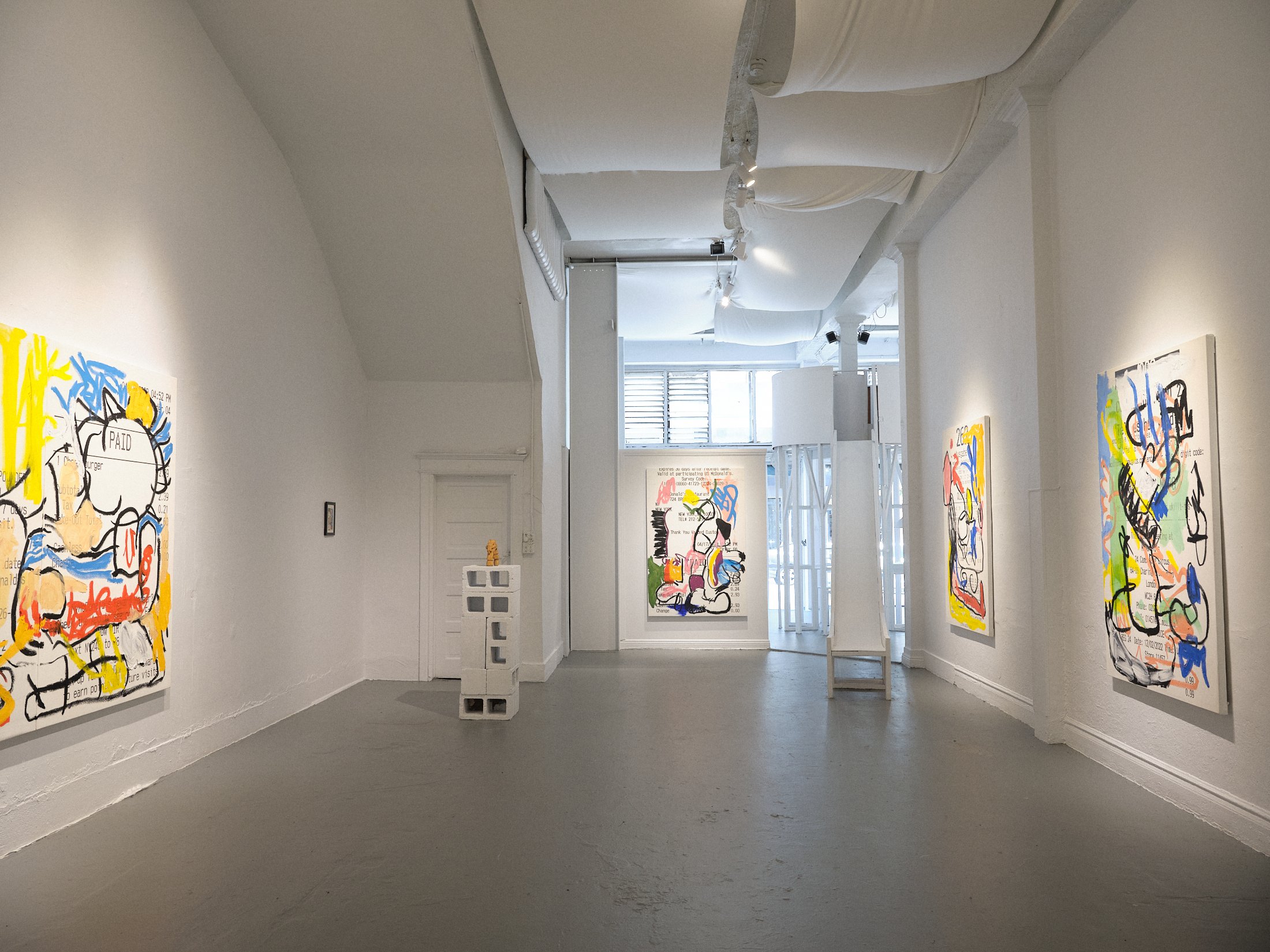 Art gallery interior with abstract paintings on white walls, featuring colorful graffiti-style art. The room has a minimalist design with smooth grey flooring and ceiling panels. A small sculpture is displayed on a pedestal.