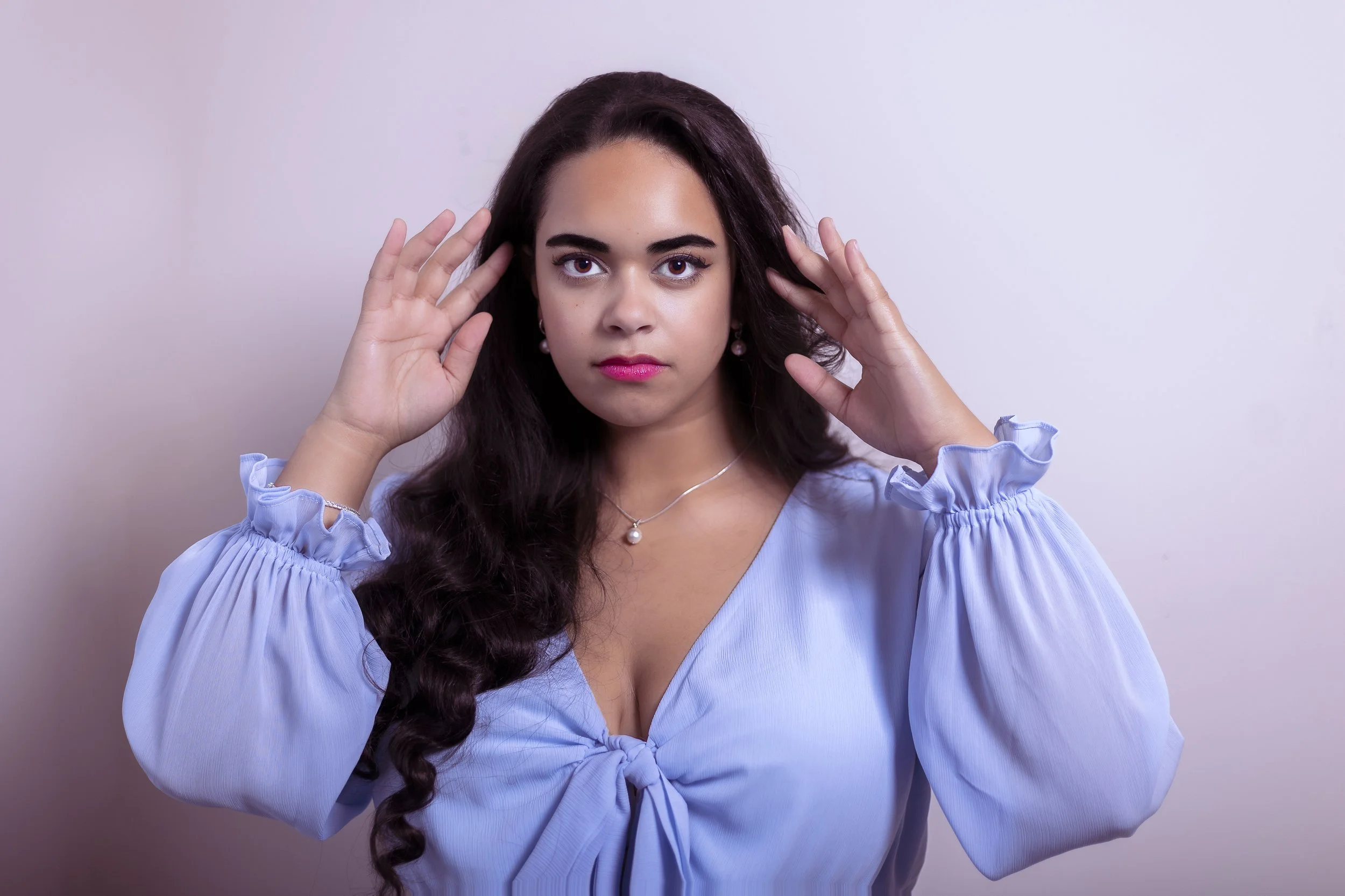 A woman with long dark hair, wearing a light blue blouse with ruffled sleeves, is holding her hands near her ears while looking directly at the camera