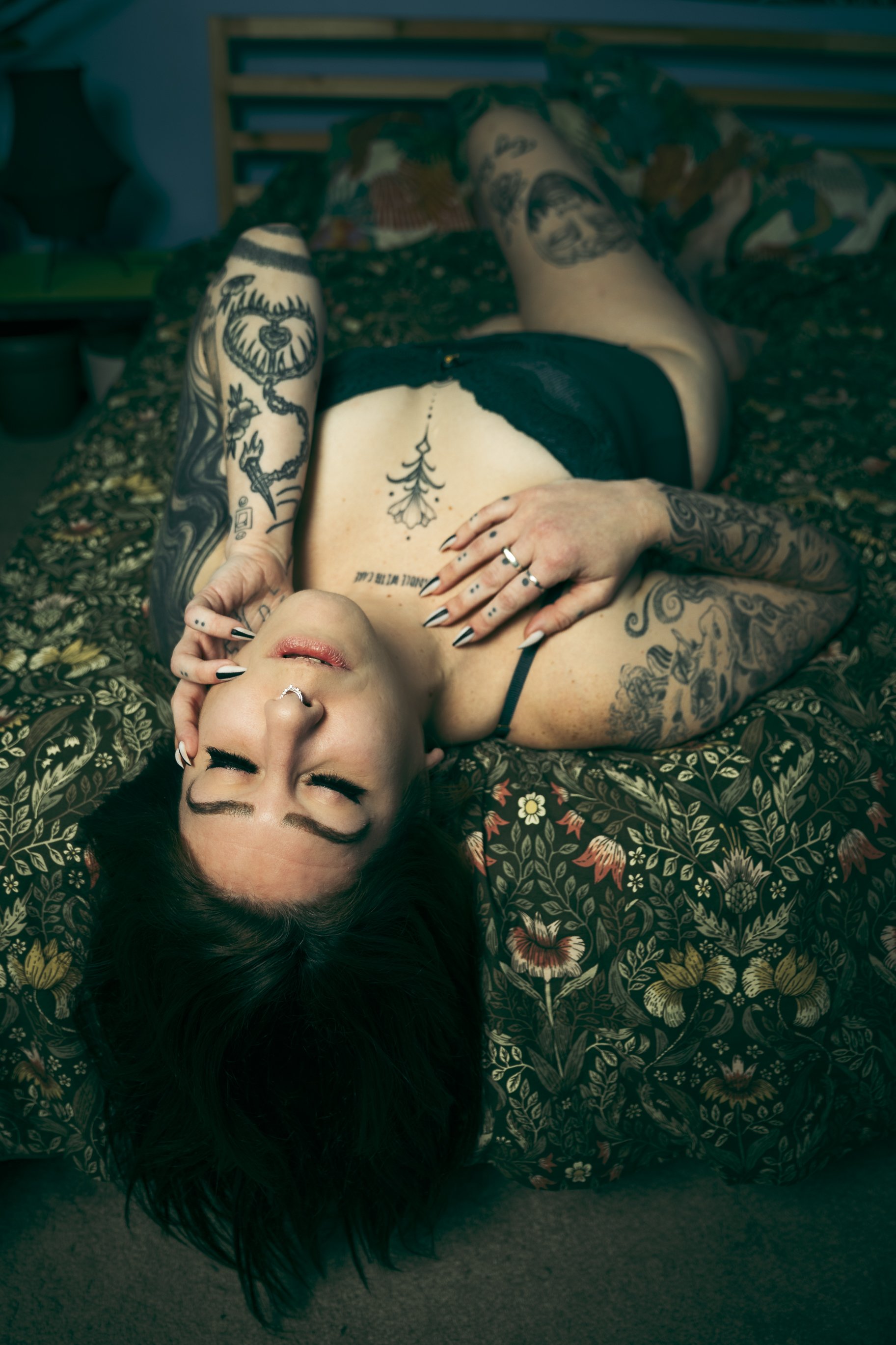 A woman with dark hair, numerous tattoos, and multiple rings lying on her back on a floral-patterned bedspread. She has her eyes closed and one hand resting on her neck and chest.