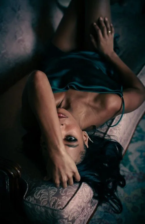 A woman lying on a vintage-style bed with dark hair, wearing a dark, silky nightdress, covering her head with her arm, in a dimly lit room.