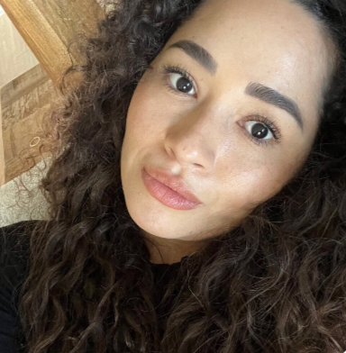 A young woman with long curly hair, natural makeup, and a slight smile, looking directly at the camera.