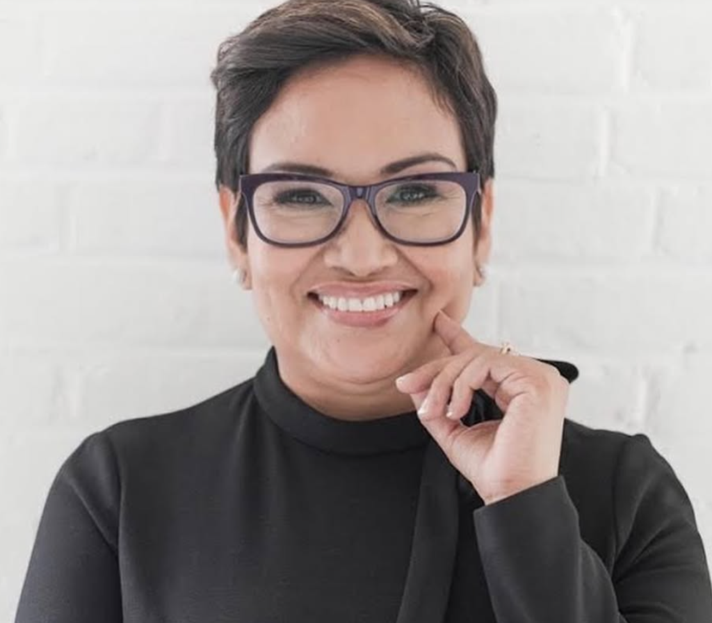 Woman with short dark hair, wearing glasses and a black top, smiling and touching her chin against a white brick wall background.