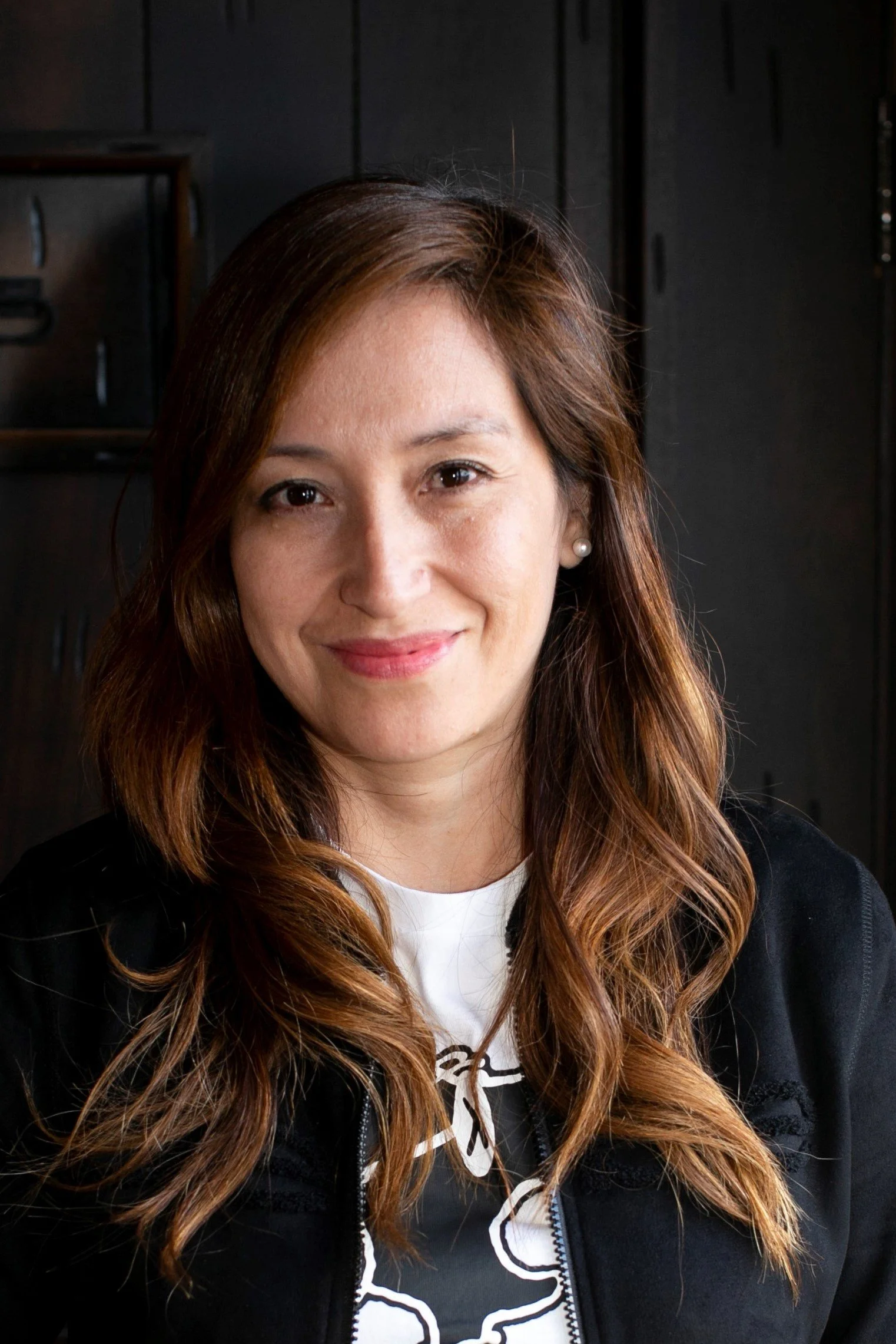 A woman with long wavy auburn hair, wearing a black jacket and white top, smiling indoors against a dark wooden background.
