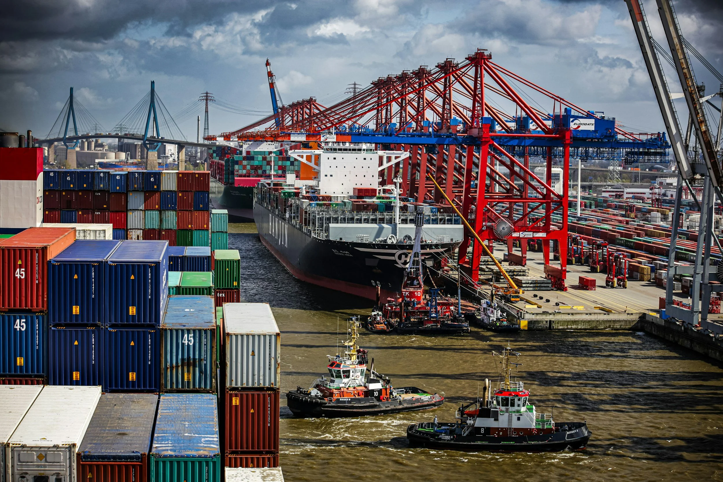 Cargo ships and tugboats docked at a busy port with container stacks and large cranes under a cloudy sky.
