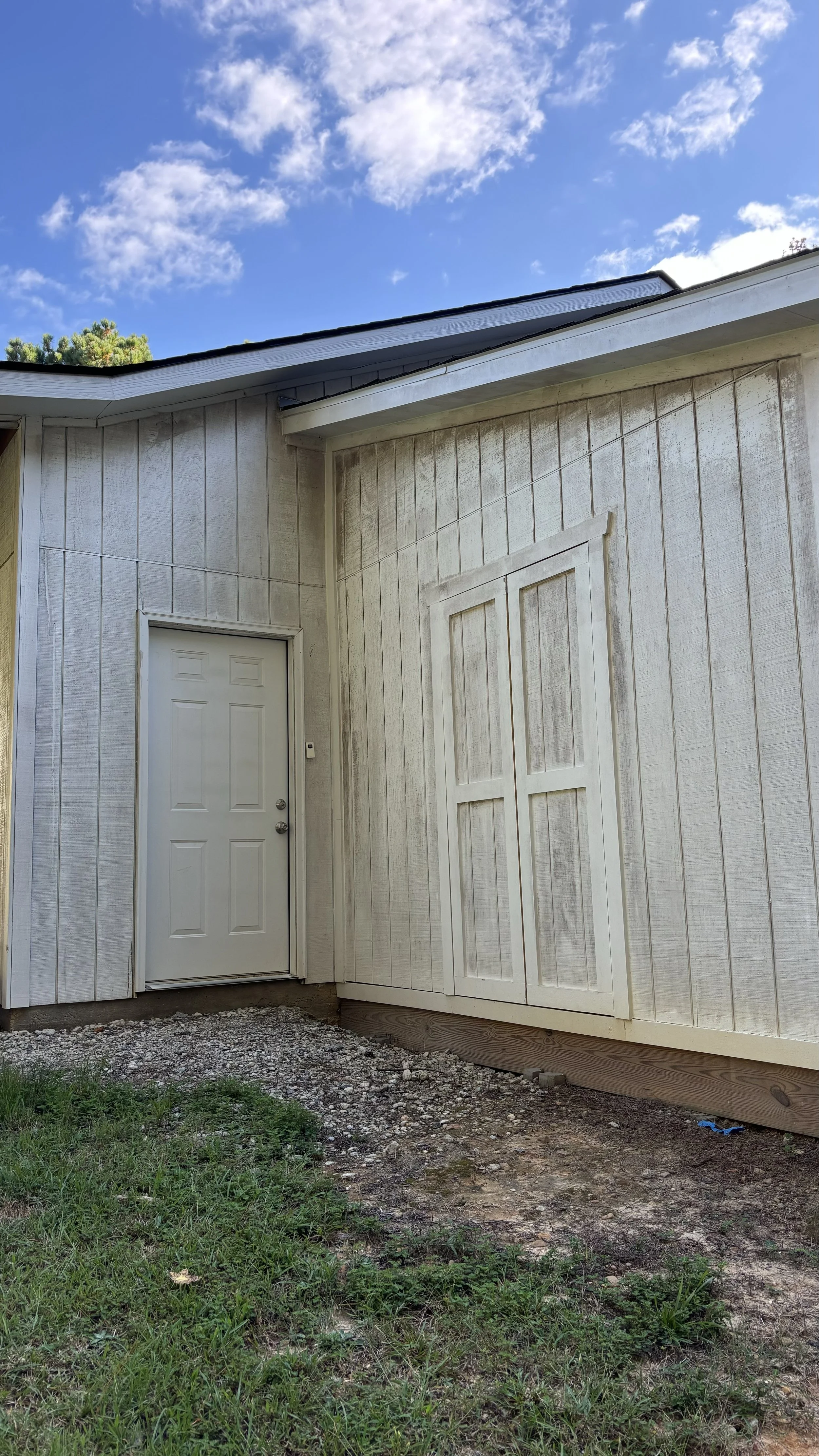 White siding building with a closed door and window, gravel and grass ground, under a blue sky with clouds.