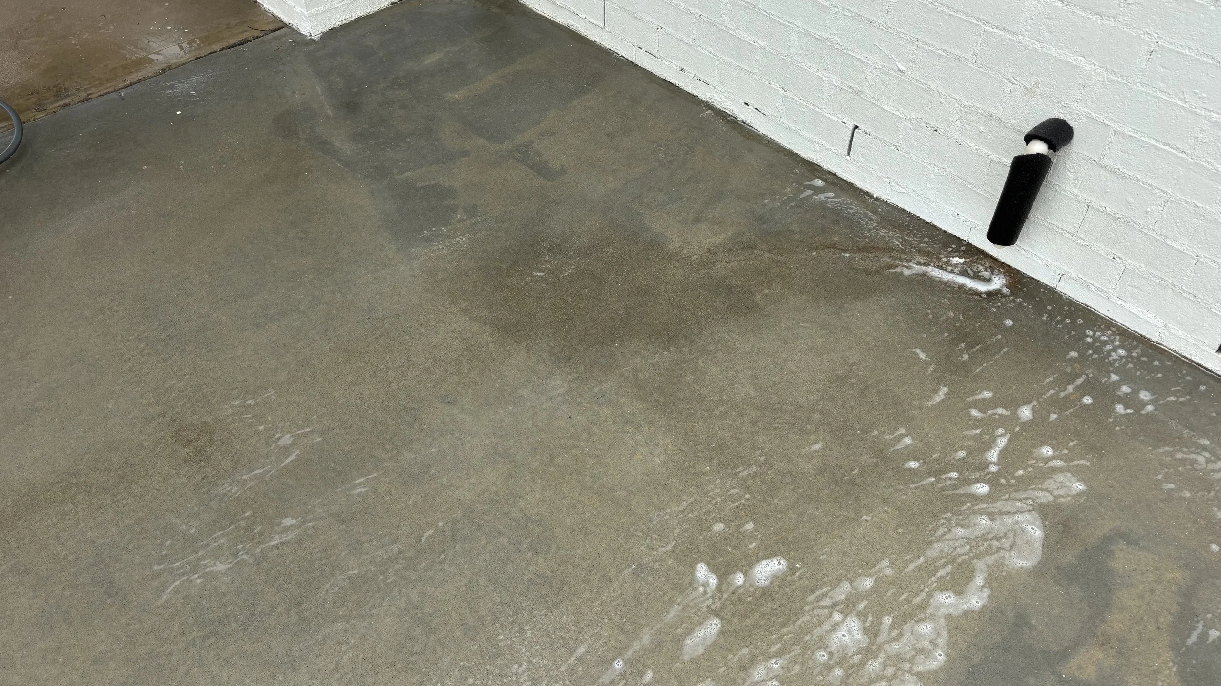 Wet concrete floor near a white brick wall with a black pipe protruding and some soap suds on the surface.