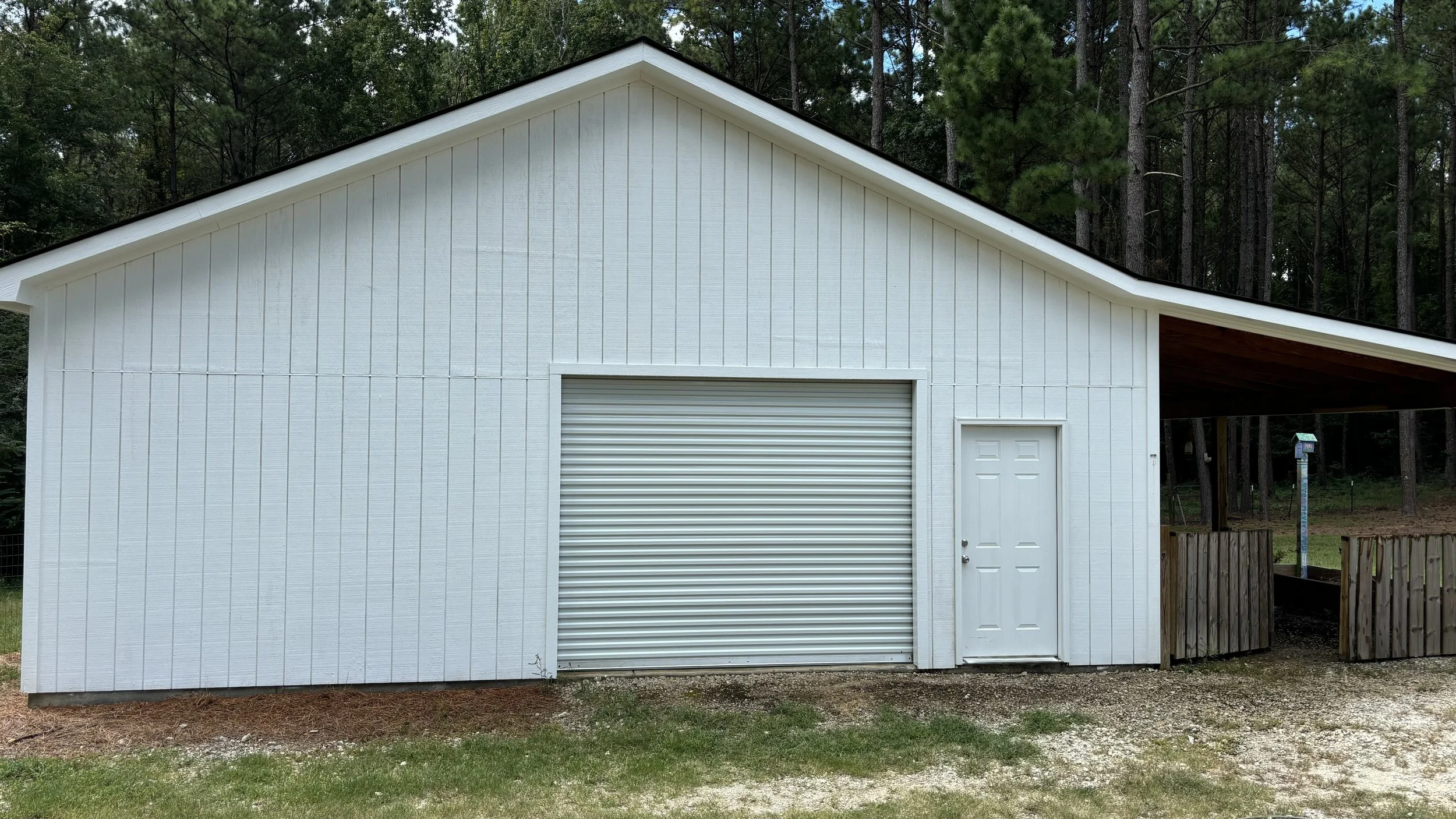White barn-style building with a metal roll-up garage door and a side entrance door, surrounded by trees.