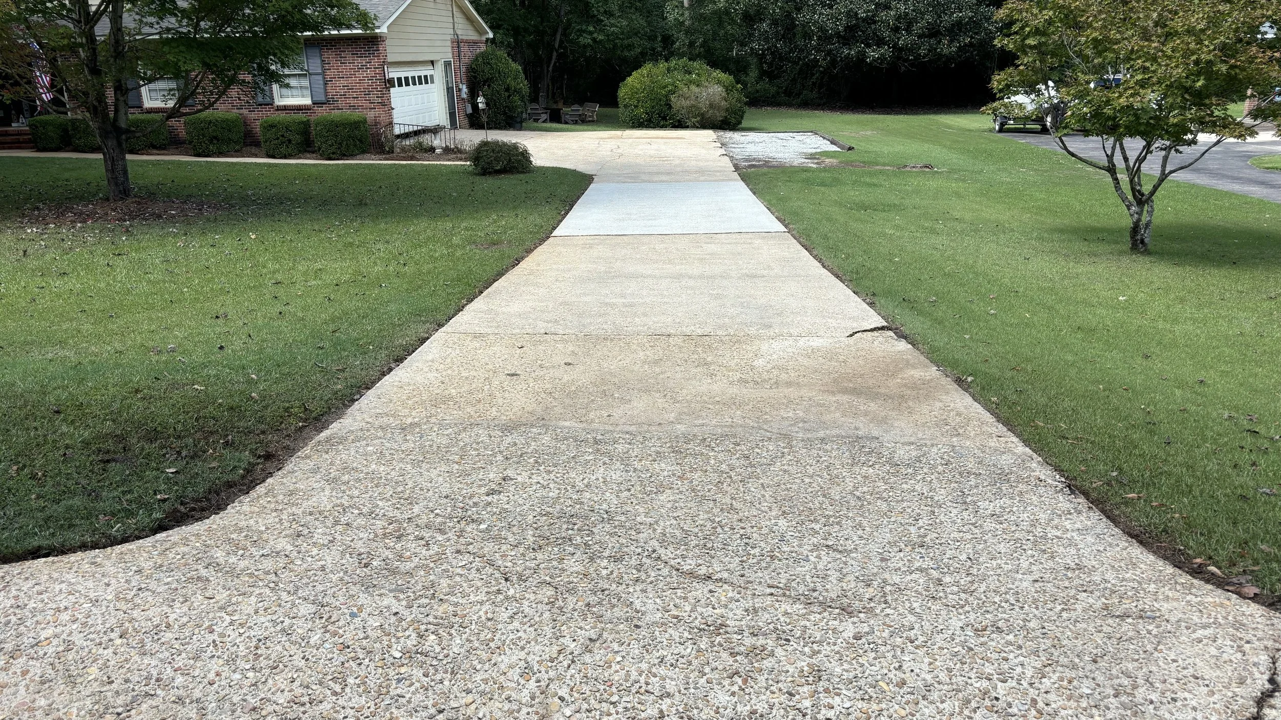 Concrete driveway leading to a suburban house, surrounded by green grass and shrubs, with a tree on the right.