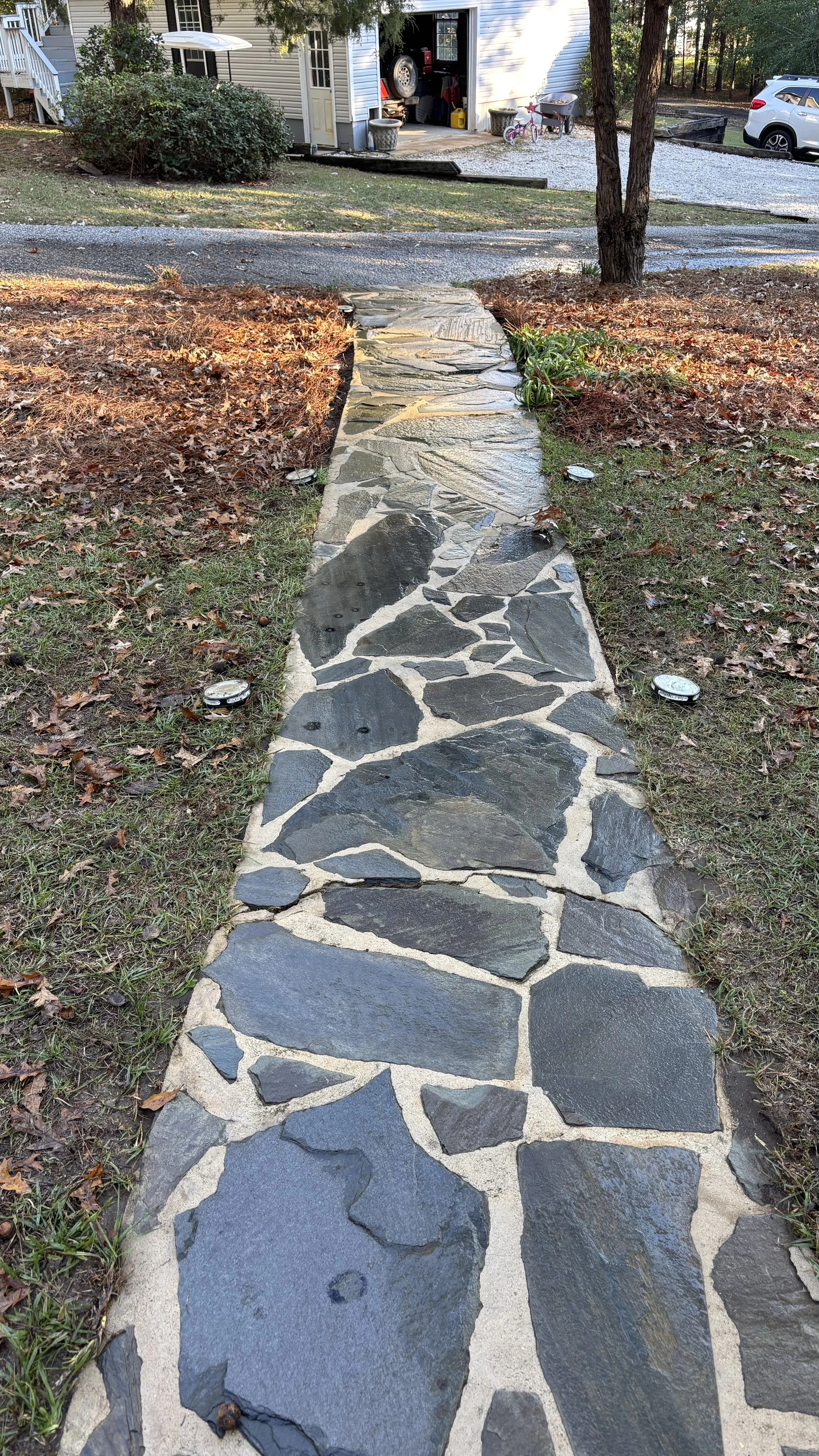 Stone walkway leading to a driveway and garage, bordered by grass and trees, with a parked car and bicycle visible.