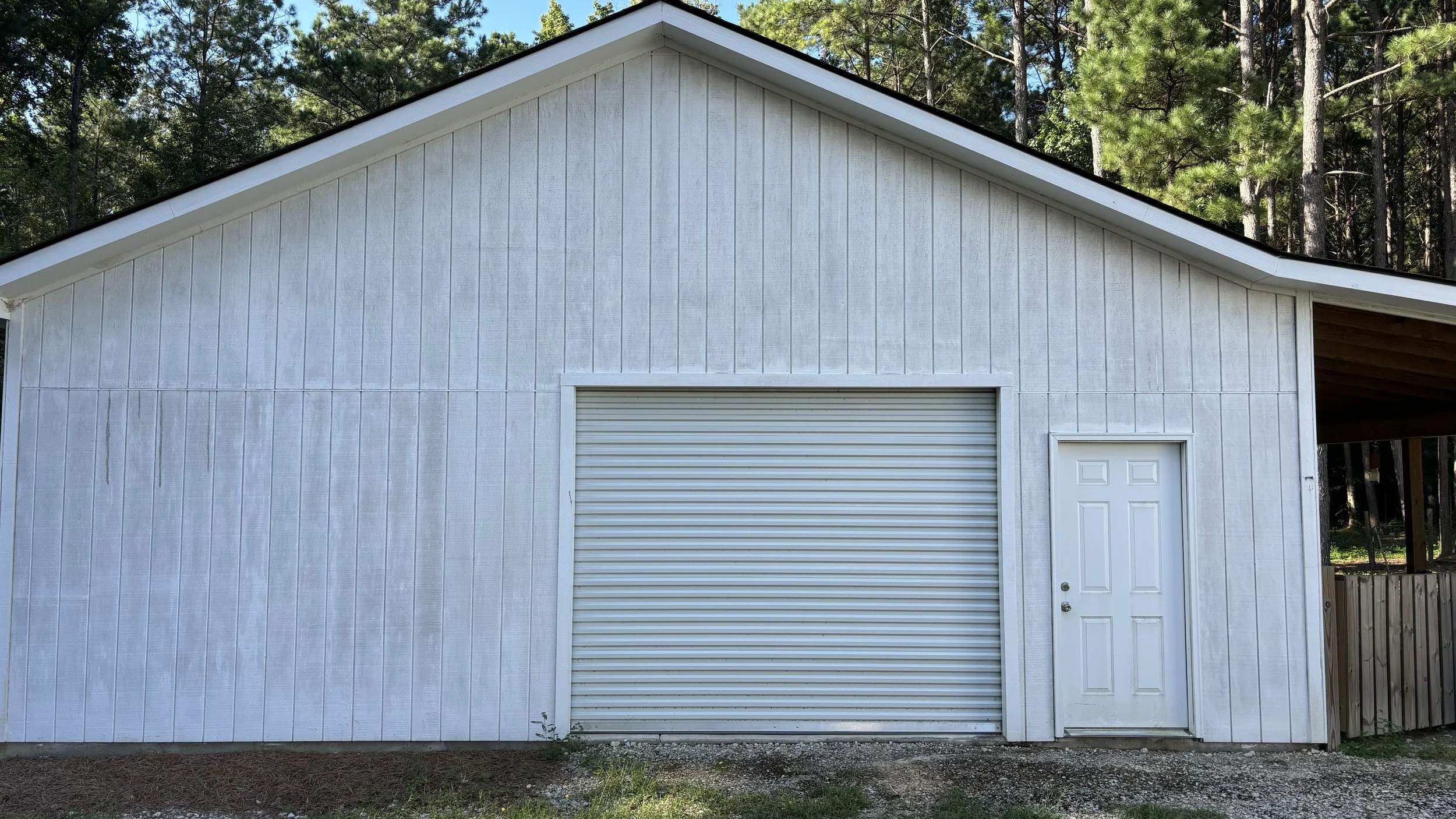 White wooden garage with a closed roll-up door and a standard entry door, surrounded by pine trees.