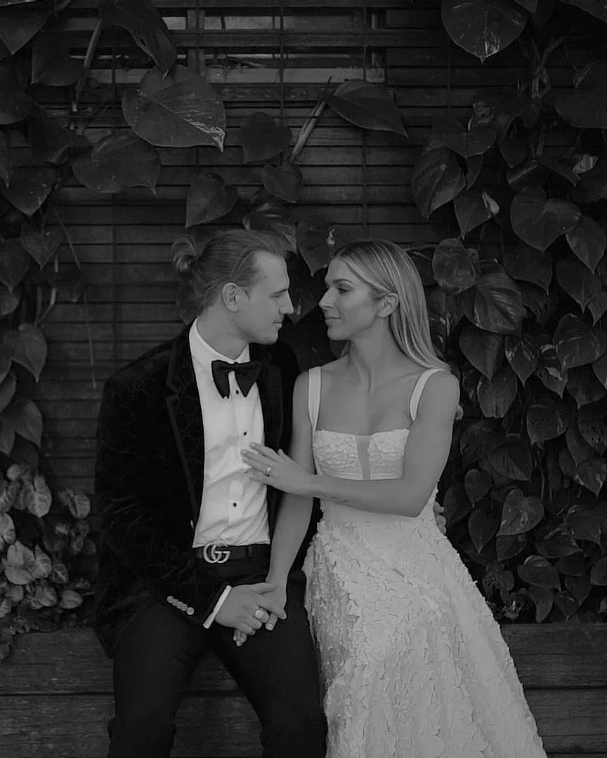 A couple in wedding attire, sitting close together with their hands clasped, surrounded by a leafy backdrop, looking into each other's eyes.