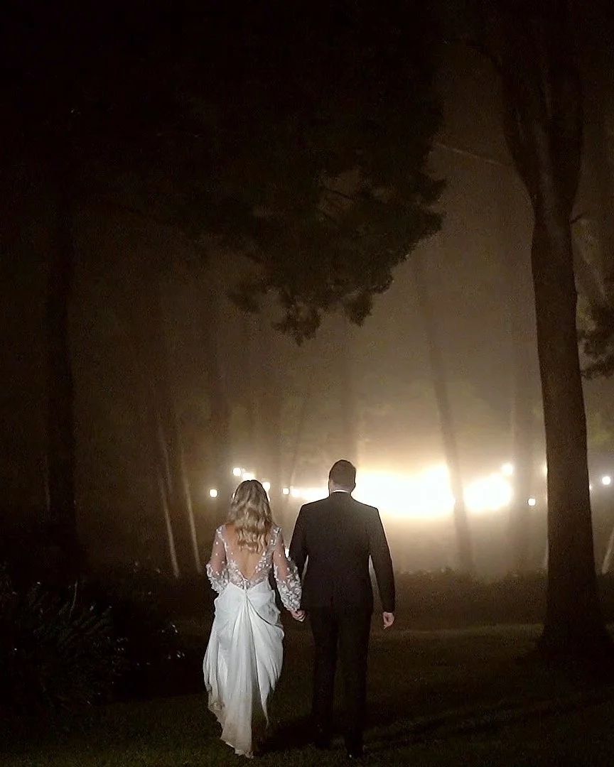 A couple in wedding attire walking hand in hand through a foggy park at night, illuminated by bright lights in the background.