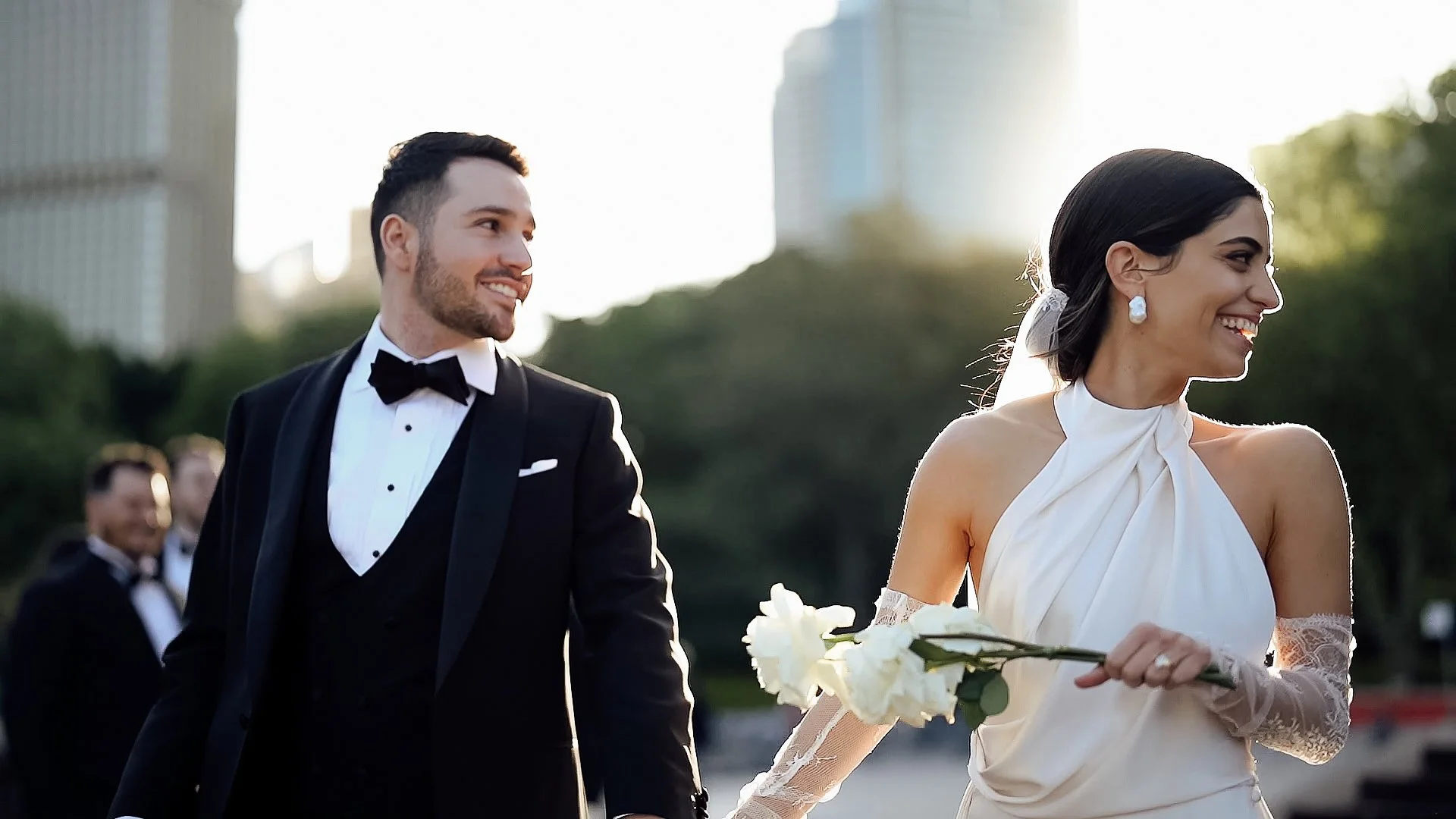A bride and groom in elegant wedding attire walking in Sydney during sunset, with the bride holding a white flower and smiling, and the groom looking at her.