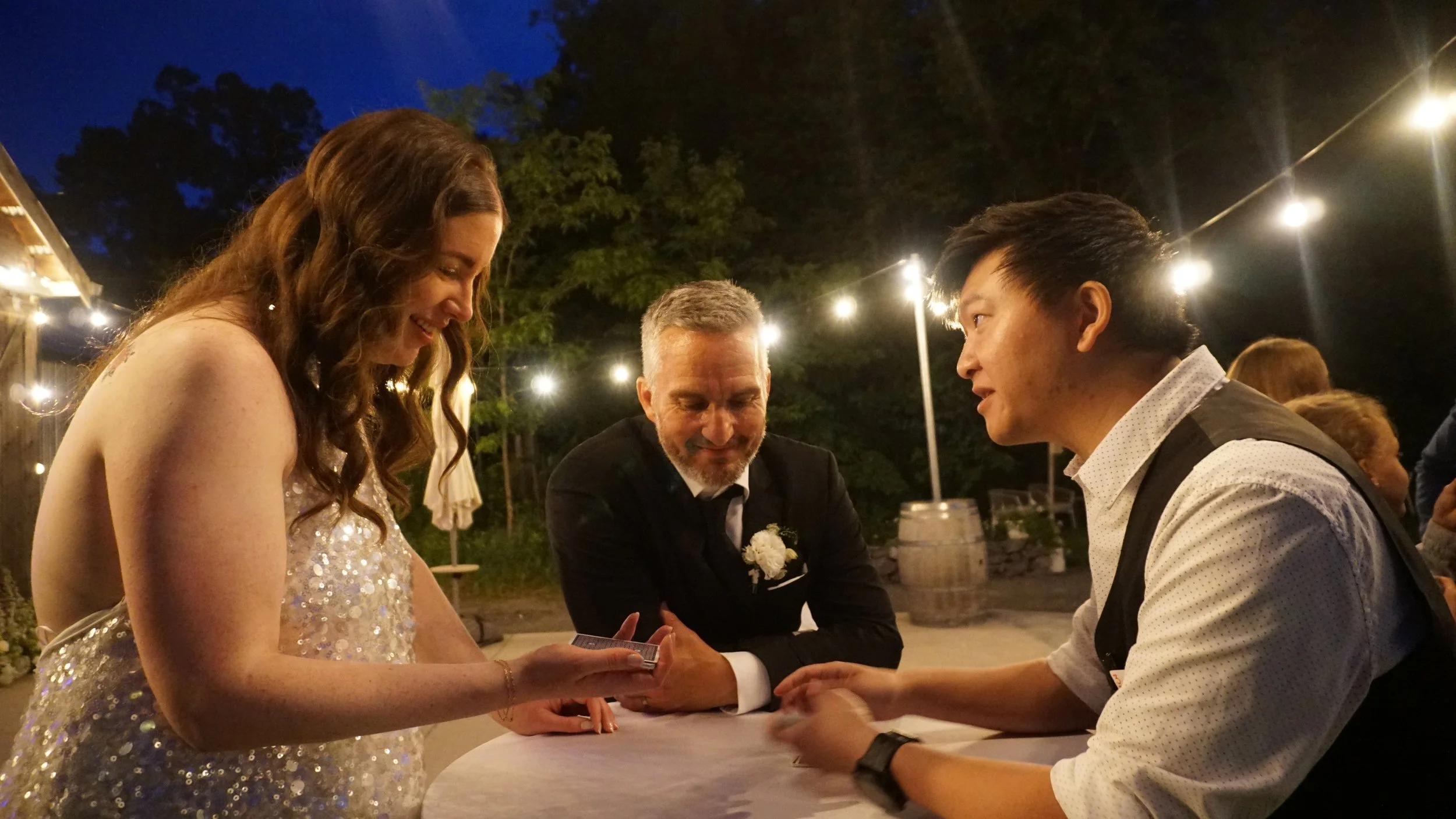 A group of four people at a wedding or formal event, sitting at a table outdoors at night. A woman in a sparkly dress is smiling and holding hands with a man in a white shirt and vests. An older man in a suit and boutonniere is also involved in the moment, and another woman with red hair is partially visible in the background. String lights illuminate the scene.