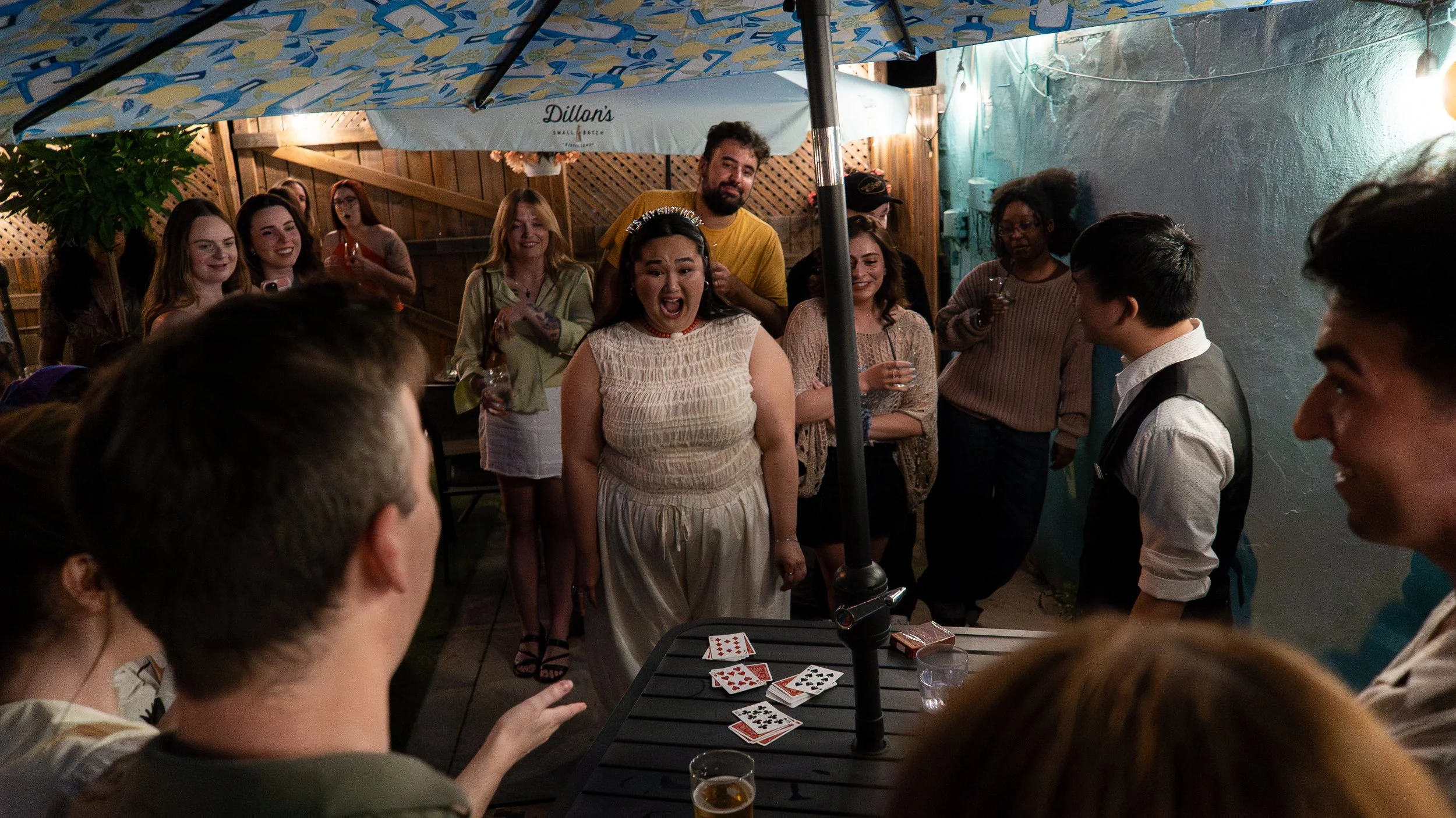 Group of people gathered outdoors at night under a large umbrella, playing cards on a table, with some holding drinks and smiling.