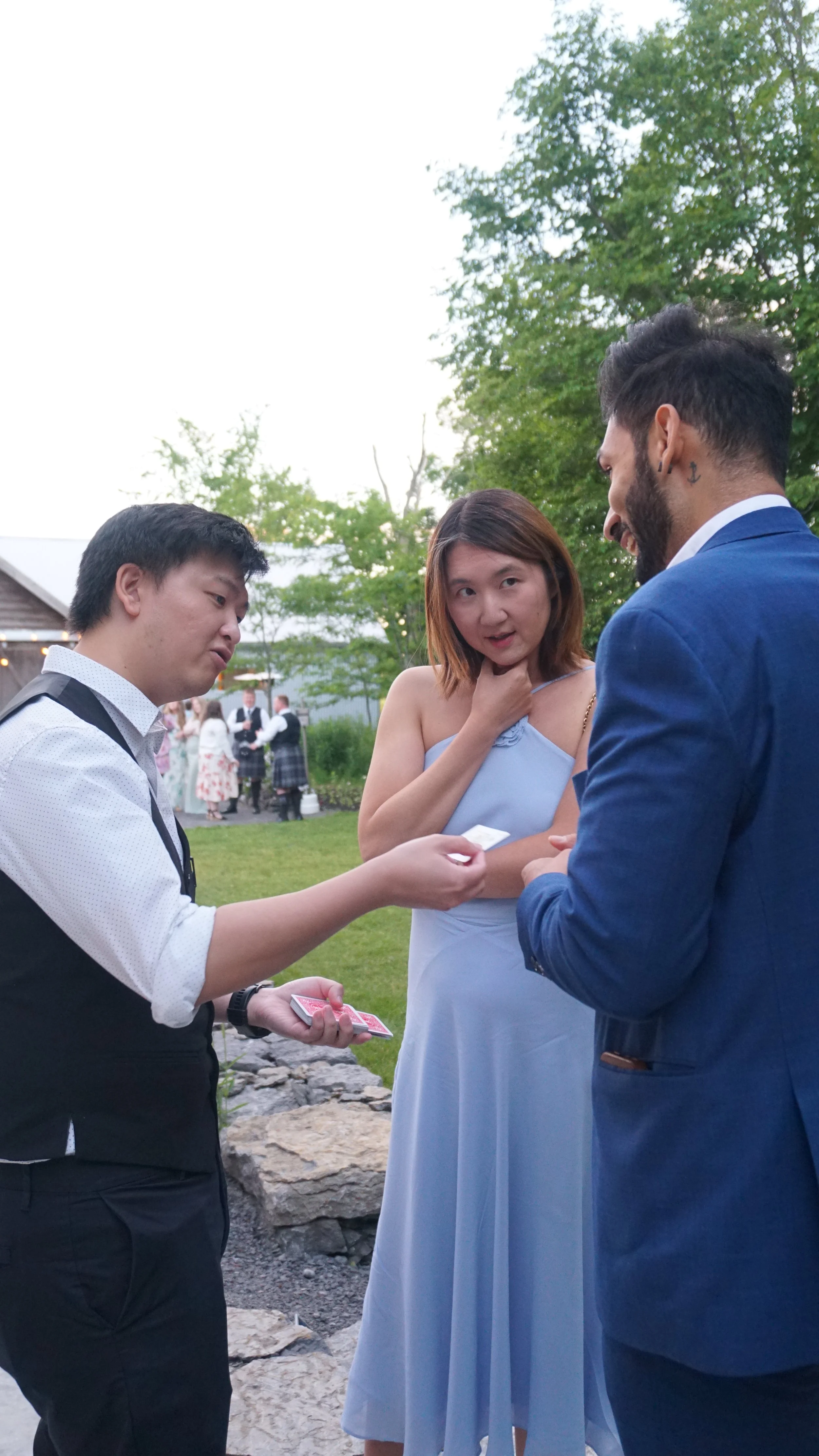A woman in a light blue dress is participating in a wedding ceremony, appearing to be the bride, as she looks at a man in a dark blue suit. A magician is performing a card trick for them. In the background, other wedding guests are visible outdoors near green trees.