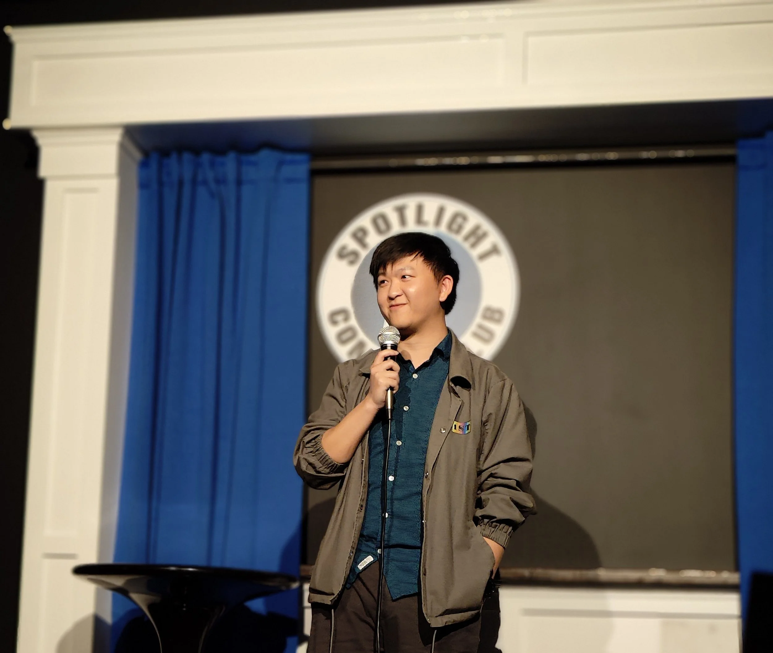 A young man standing on stage holding a microphone during a comedy show at Spotlight Comedy Club. He is wearing a gray jacket over a dark blue shirt and has a thoughtful expression. Blue curtains and the club's logo are visible in the background.