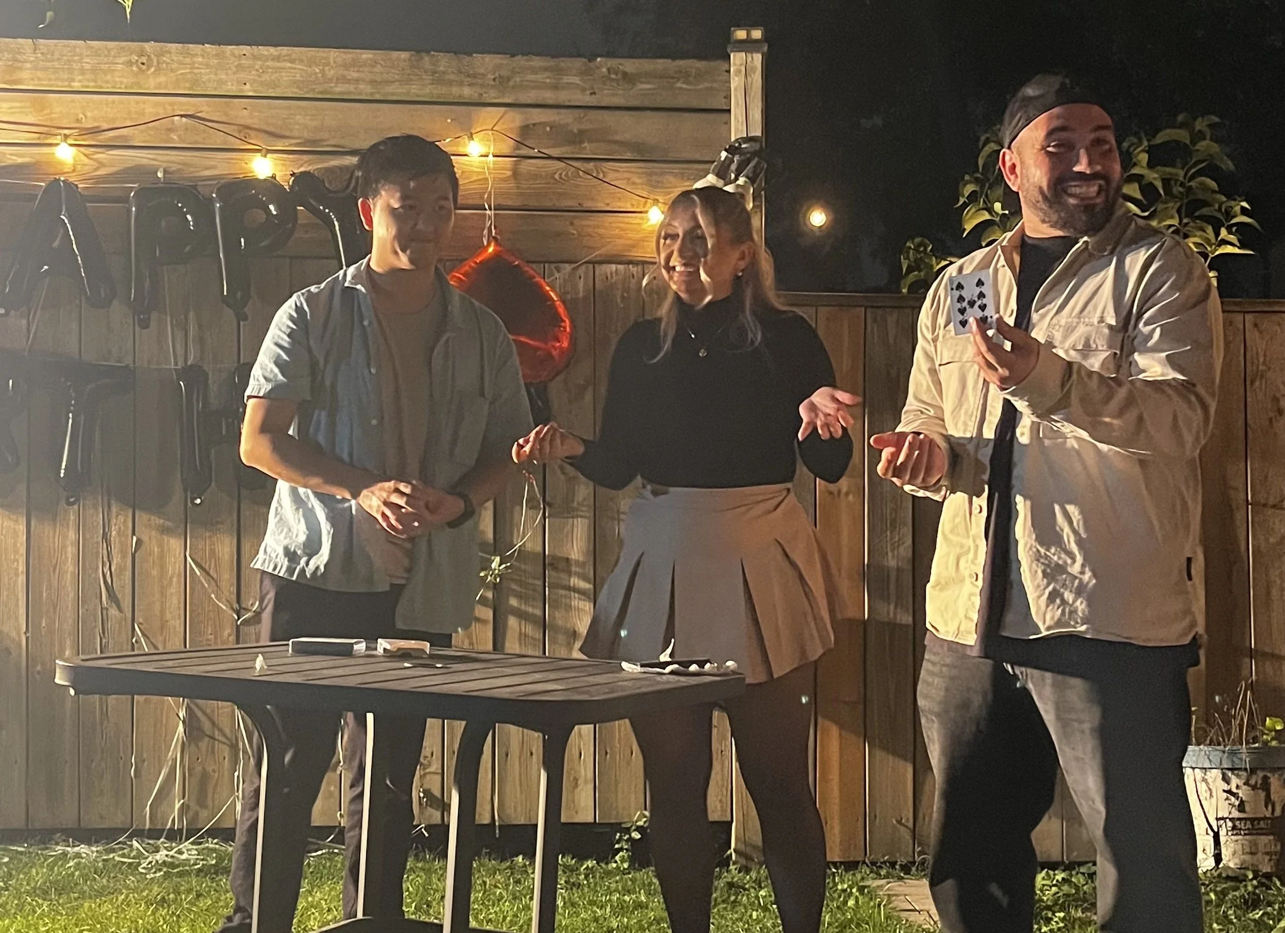 Three people standing outside in a backyard at night, with a wooden fence and string lights in the background. One person on the right is holding a playing card, smiling. The woman in the middle is talking, and the man on the left is standing with his hands clasped.