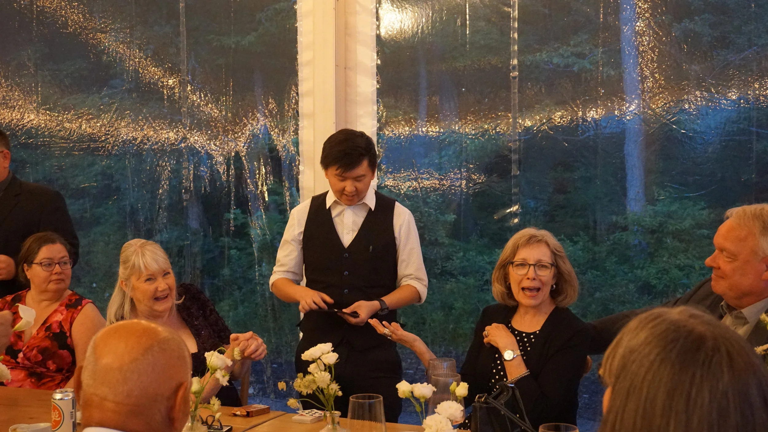 A group of people sitting around a dining table at an outdoor-themed restaurant, with a transparent plastic wall reflecting trees and lights, and a young waiter standing in the middle serving the table.