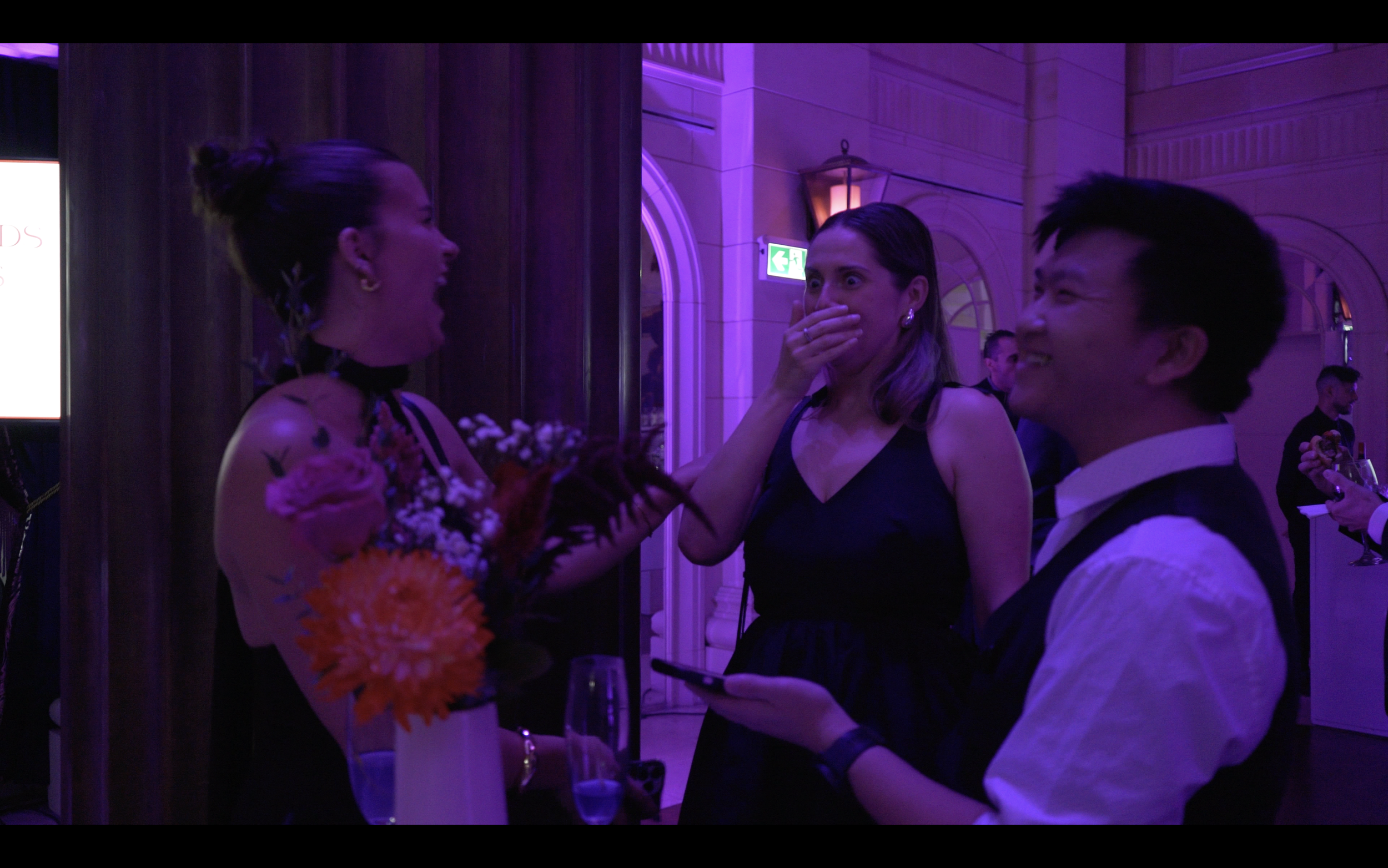 Three people at a social event, two women and one man, are laughing and smiling in a warmly lit room with purple lighting. The woman on the left holds a bouquet of flowers. The woman in the middle covers her mouth with her hand, and the man on the ri