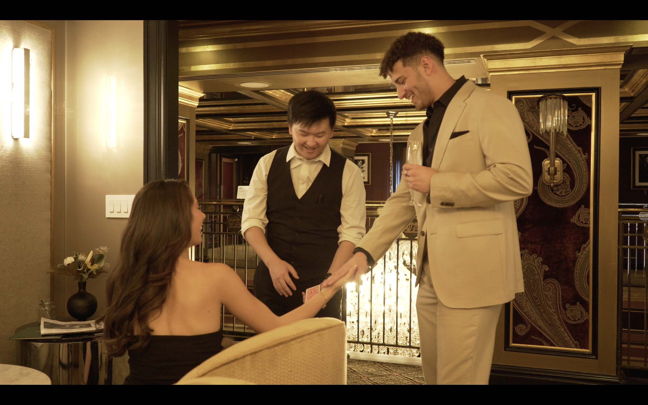 A woman sitting at a table with long brown hair wearing a strapless black dress, receiving a ring from a man in a beige suit holding a glass of wine, with a waiter standing nearby in a hotel or upscale restaurant decorated with gold accents and ornat