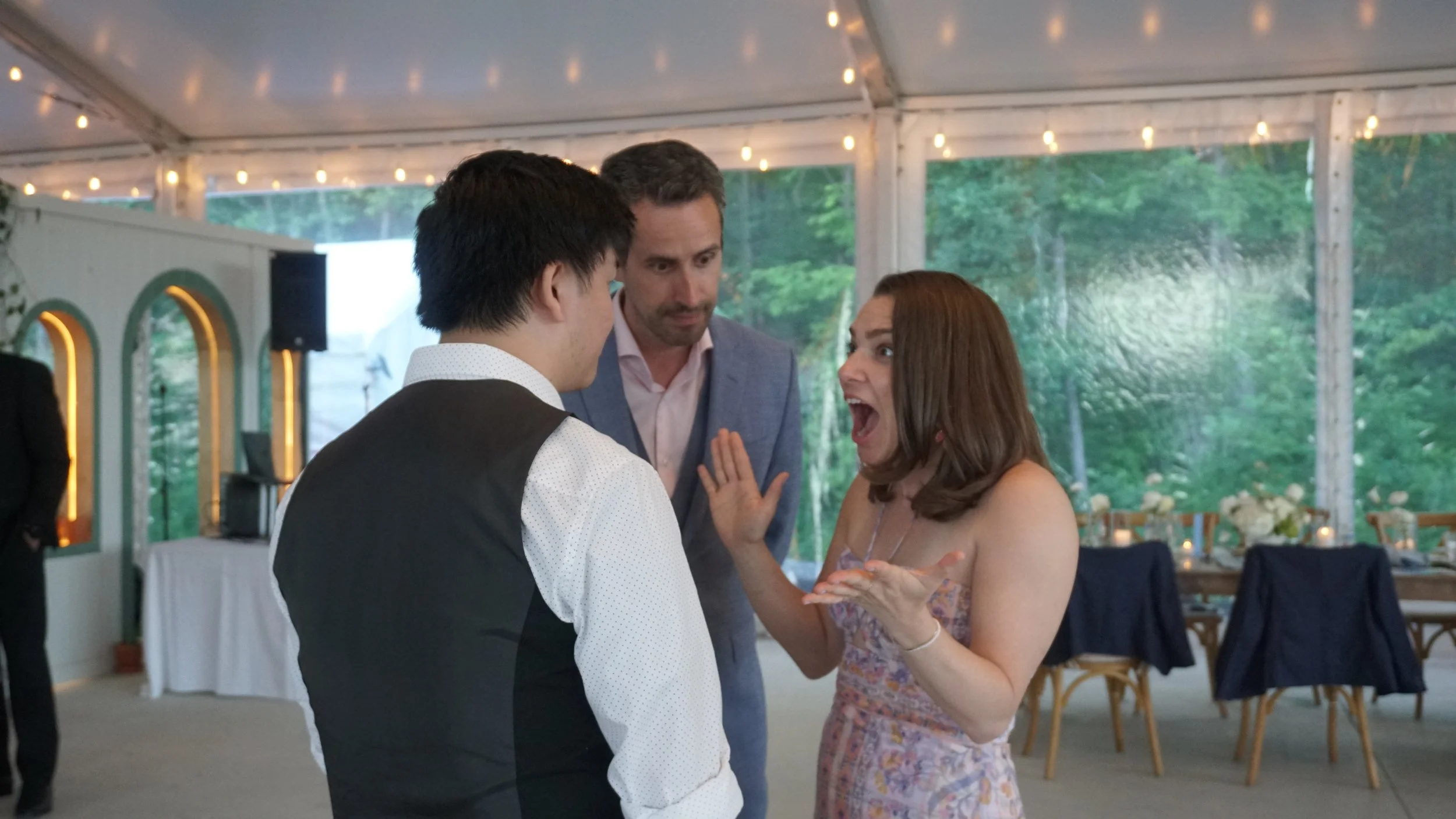 Three people at a wedding reception having a lively conversation. A woman appears to be surprised or excited, while two men listen intently.