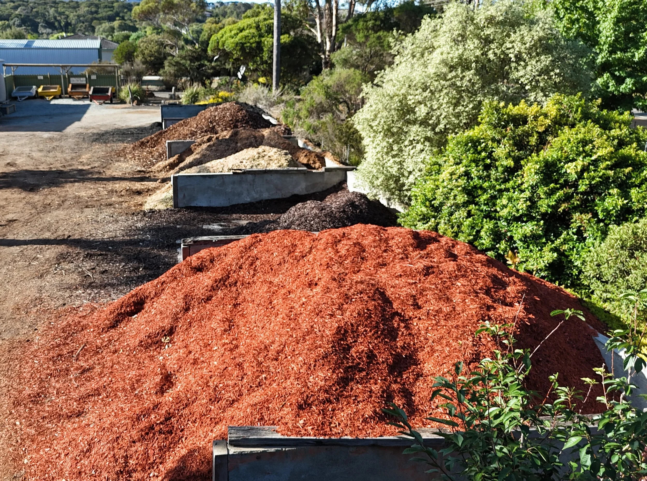 Several piles of mulch in different colours and other soils and sands are lined up outdoors next to green bushes and trees.