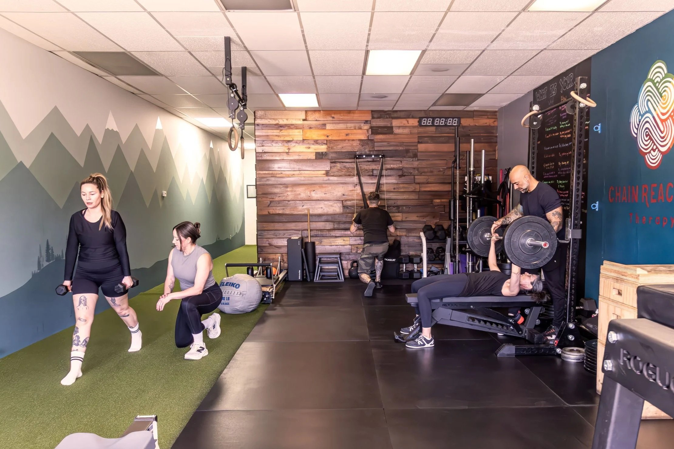 People exercising in a gym, including a woman on a bench, a woman stretching on the floor, and another stretching on the black mat, with gym equipment like weights and treadmills around.