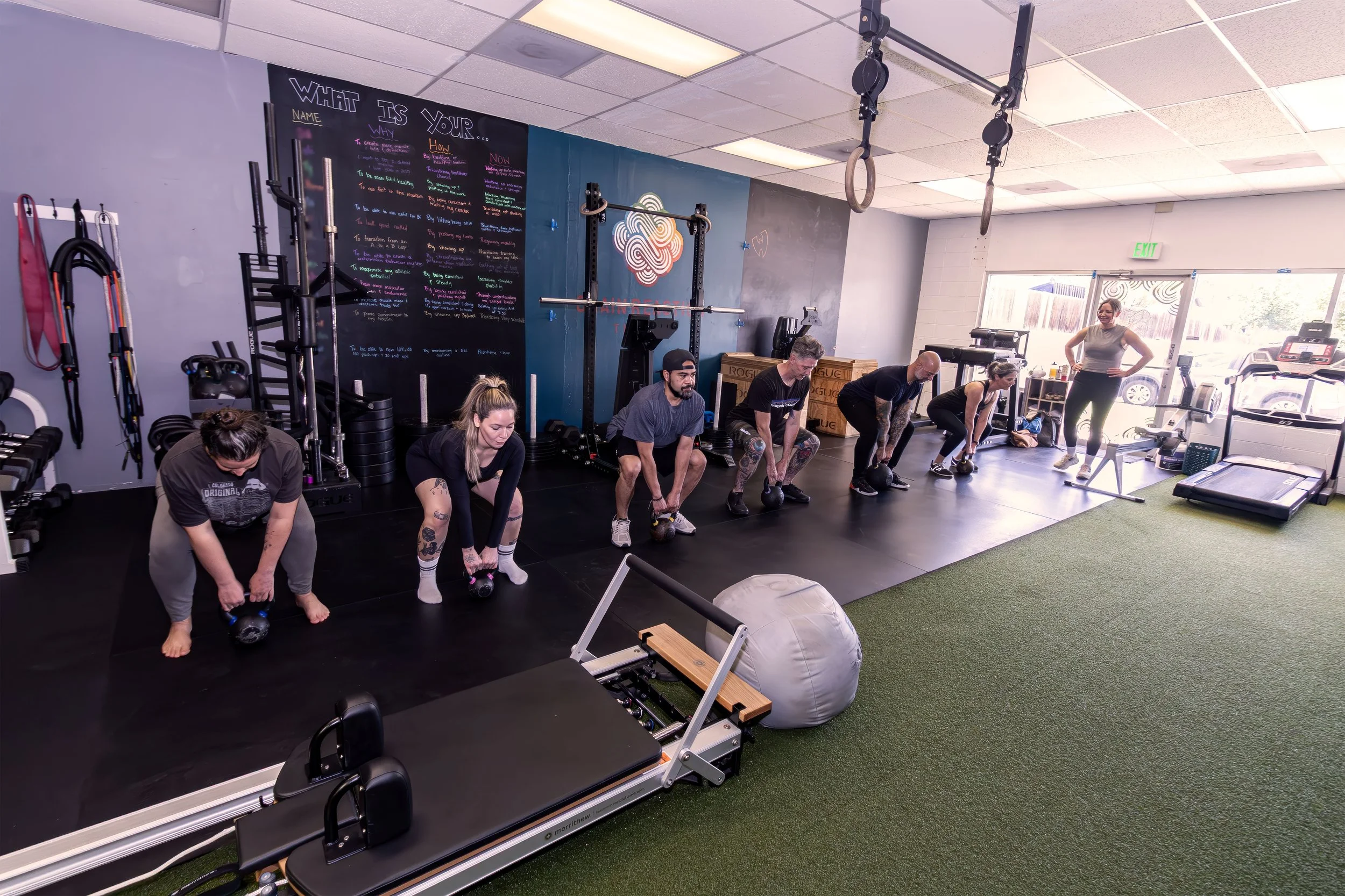 People working out in a gym with weights and exercise equipment, including a person lying on the floor with legs raised, two people standing and lifting weights, and a woman doing a step on a small platform, inside a fitness studio with a blue and wooden wall and blackboards with writing.