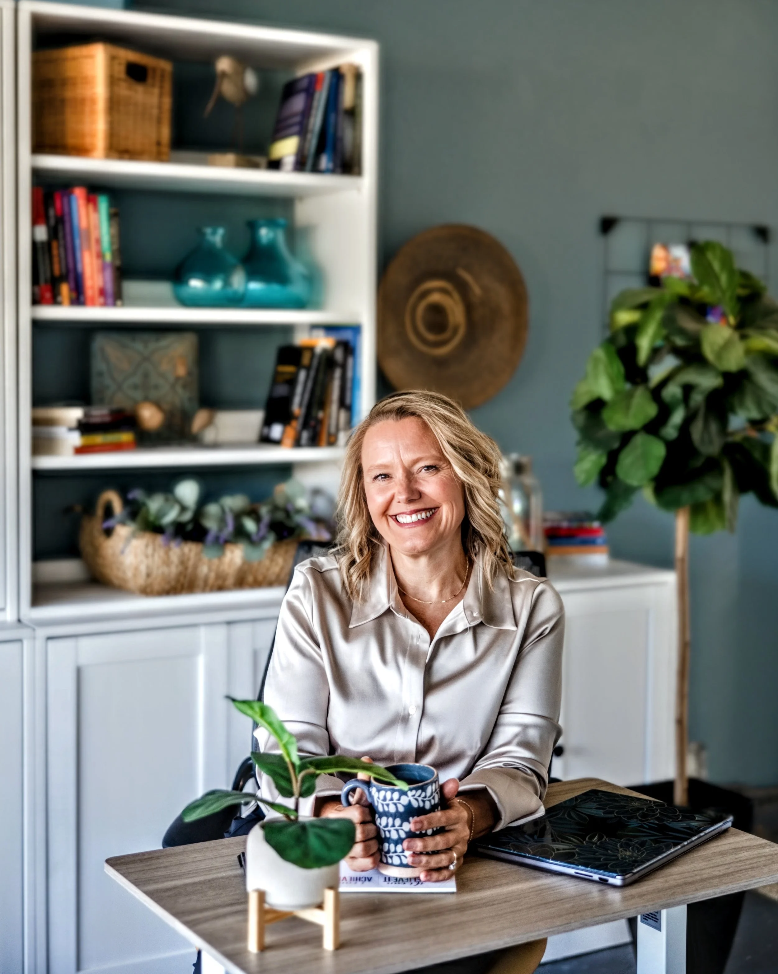 Whitney at her desk with a coffee mug in her hand and a bookcase behind her. She is smiling towards the camera.
