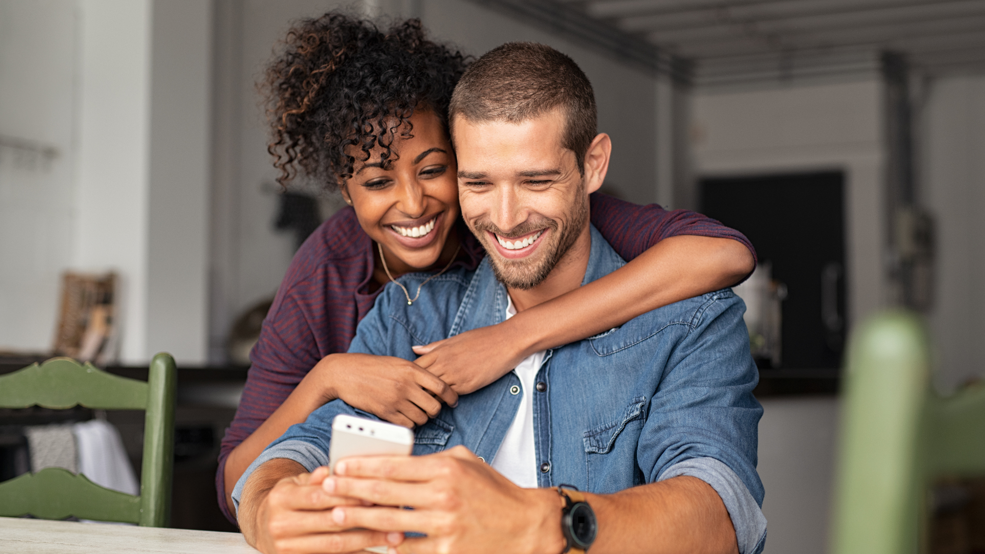 Man smiling at his phone at home, with his wife looking over his shoulder and happy at what she sees, too
