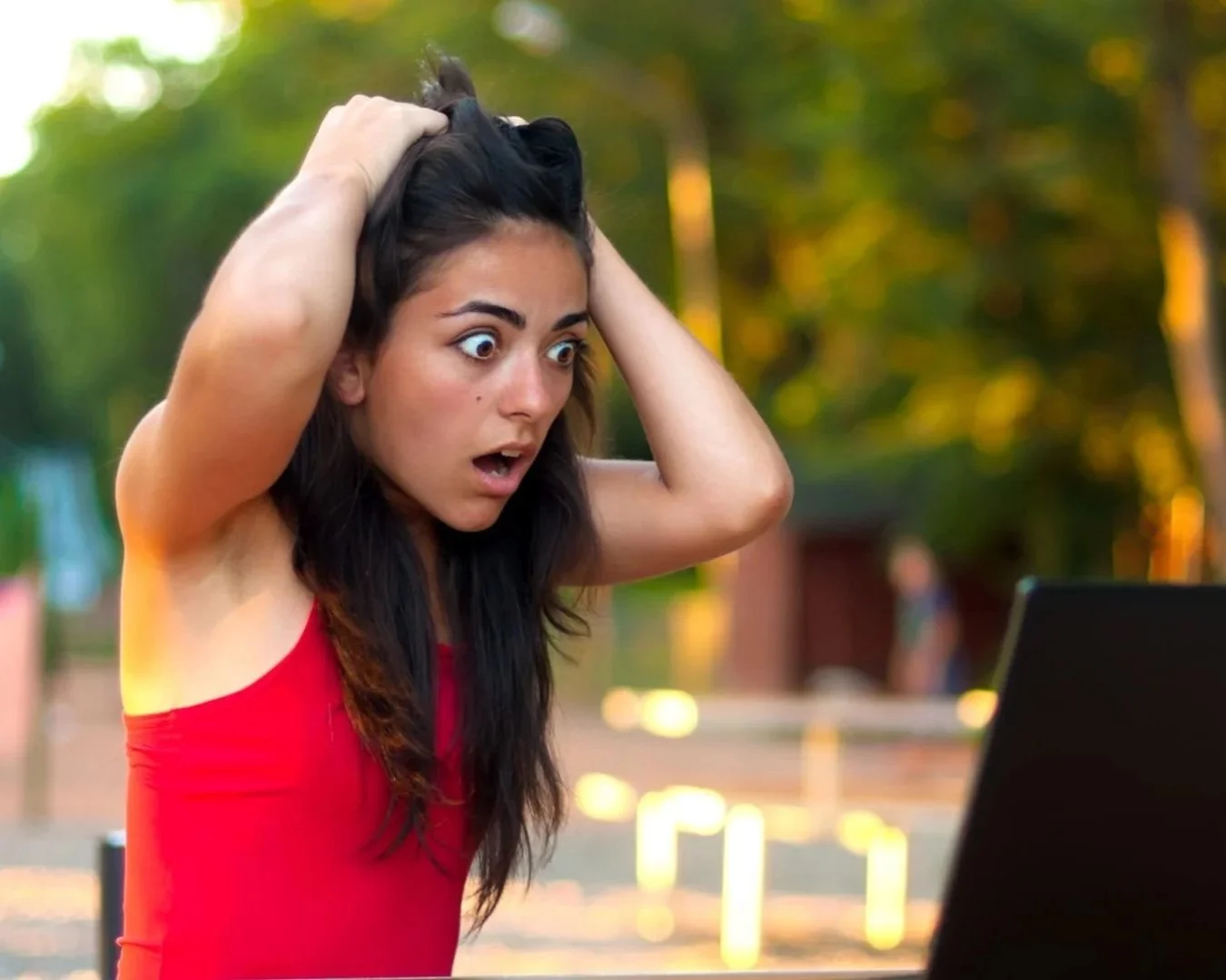 Woman in a red tank top looking aghast at her open laptop outside with a park setting in the background
