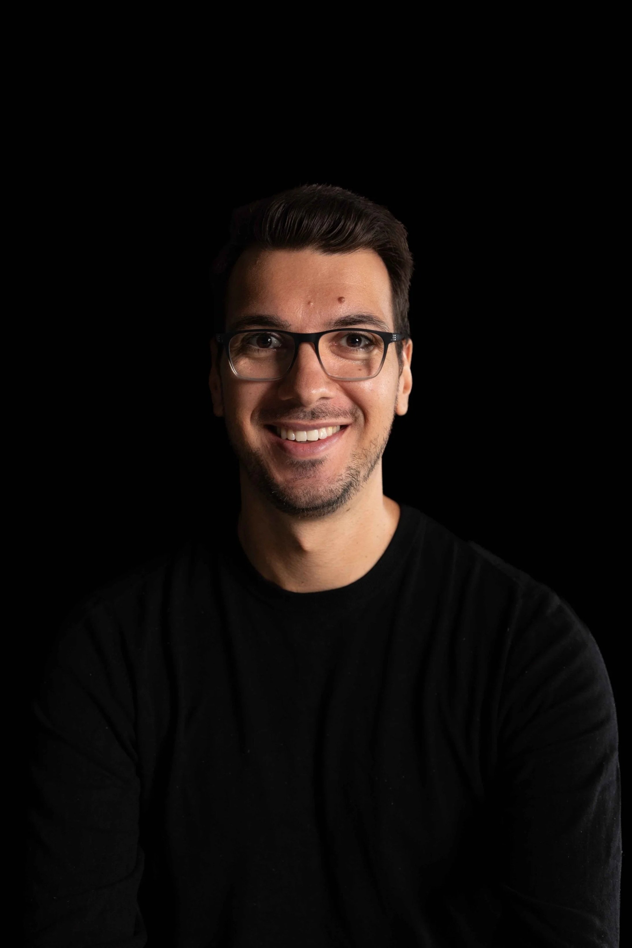 A smiling man wearing glasses and a black shirt, posing against a black background.