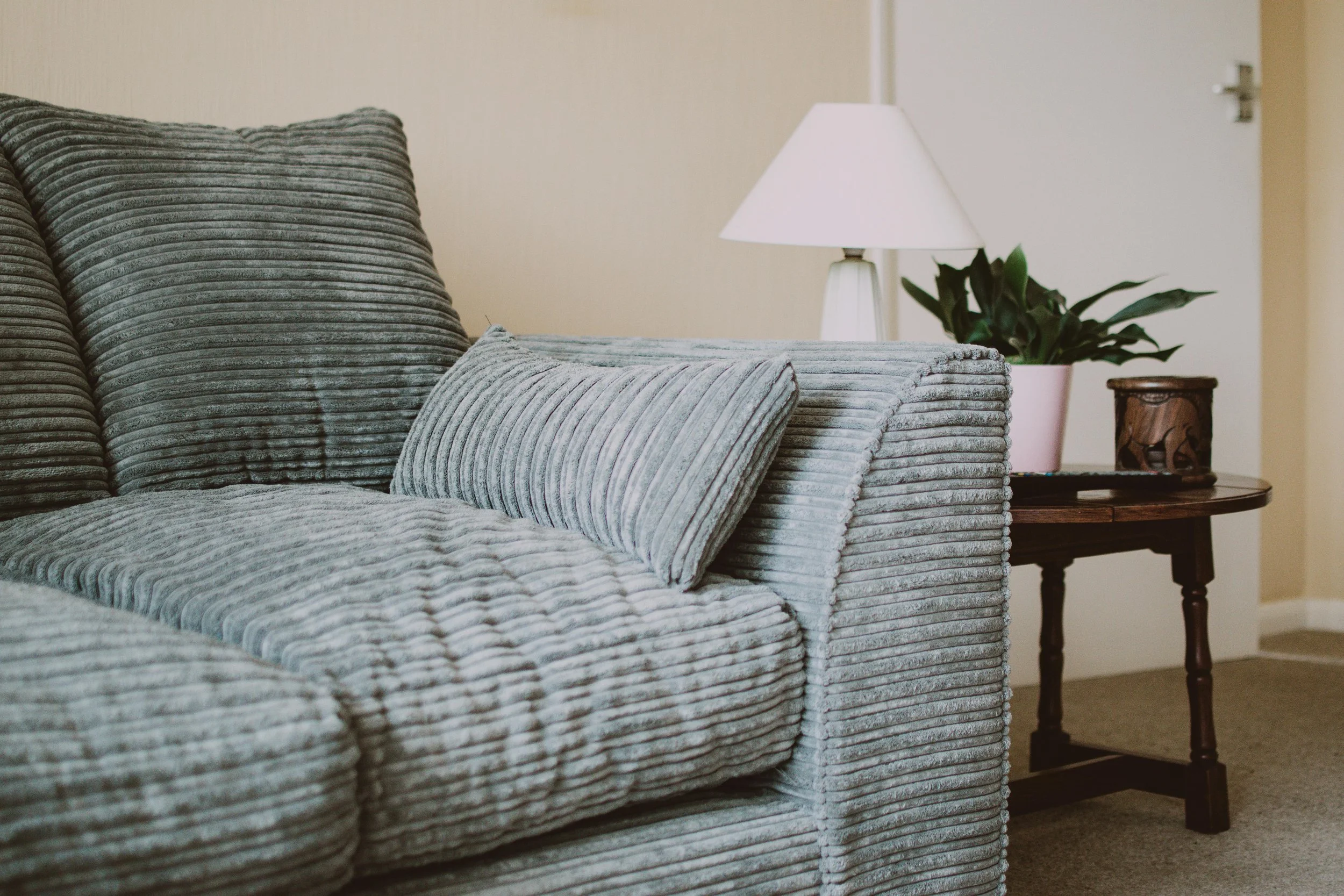 Gray corduroy sofa with cushions next to a side table holding a lamp and potted plant.